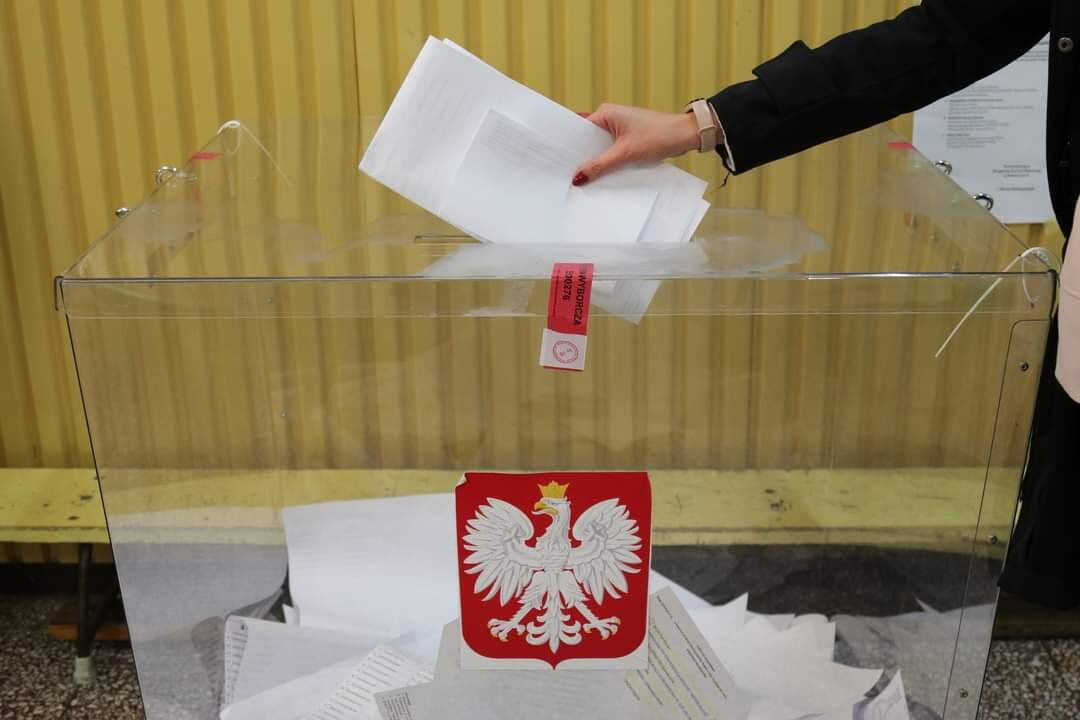Ręka wkładająca kartki do przezroczystej urny z godłem Polski.
