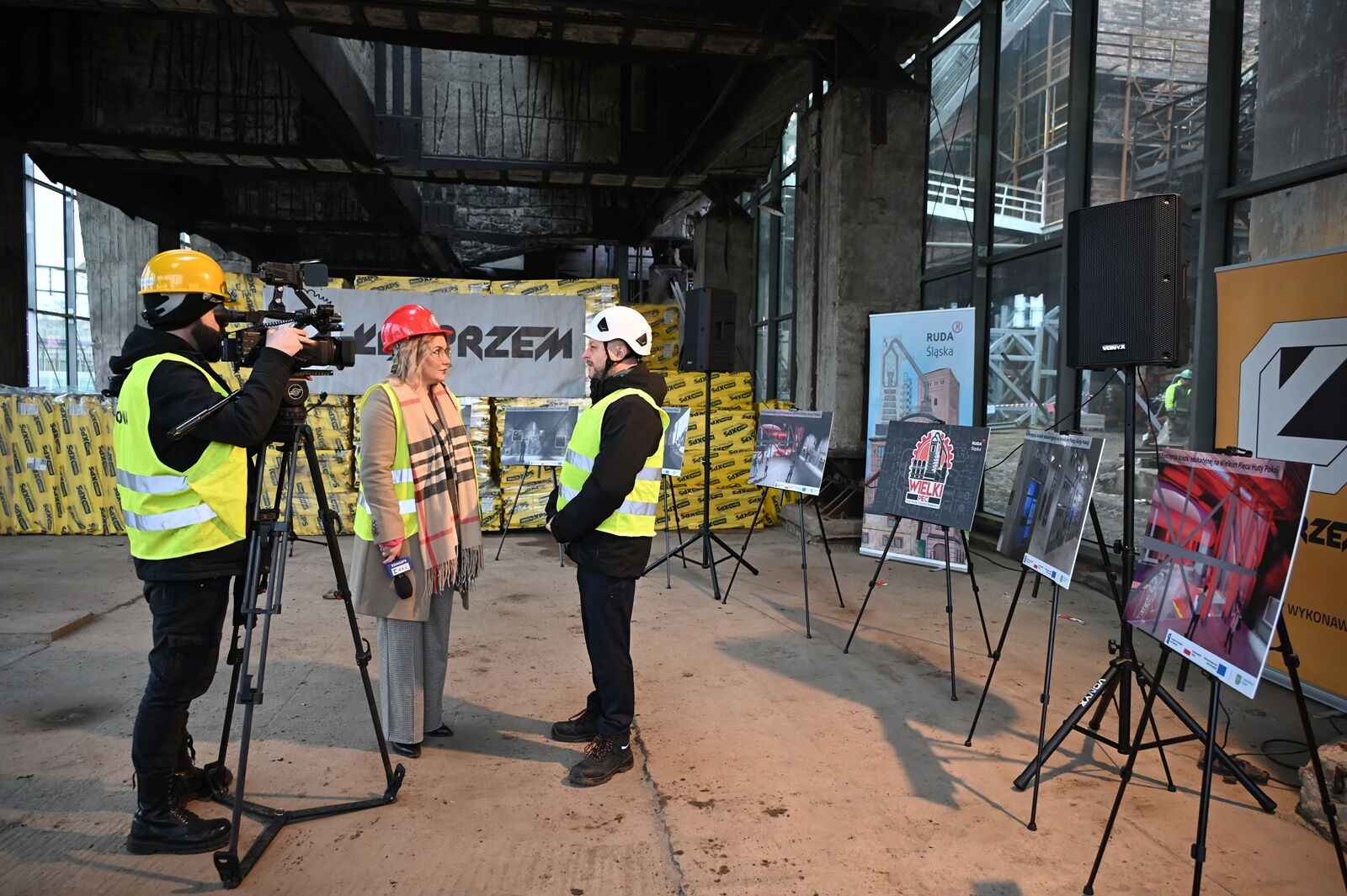 Trzy osoby: mężczyzna stoi przy kamerze, przed nią stoi inny mężczyzna, a obok niego kobieta, wszyscy w kaskach. Stoją w przestrzeni industrialnej, widać też tablice na sztalugach.