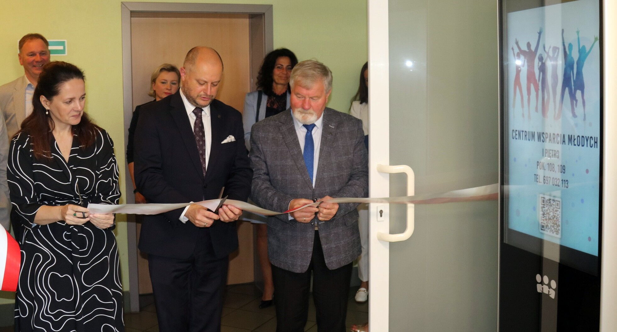 Trzy osoby przecinają wstęgę zawieszona na drzwiach, w tle cztery osoby.