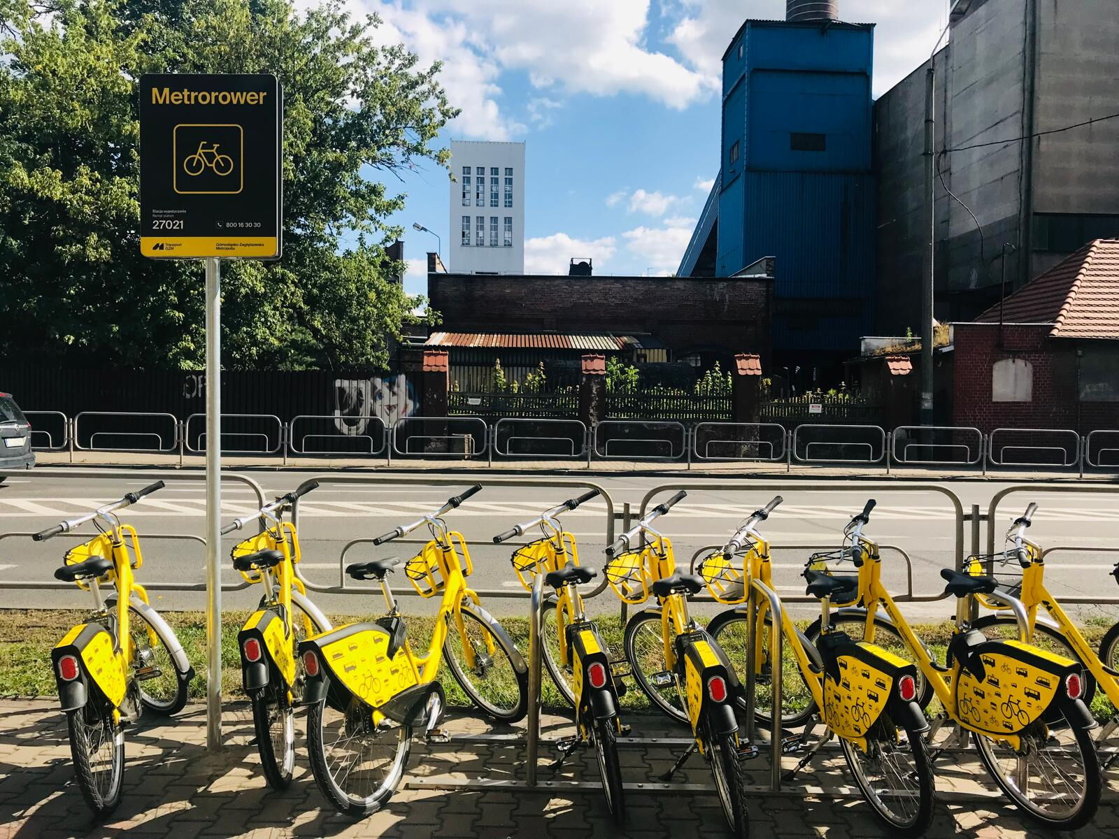 Na pierwszym planie stoi kilka żółtych rowerów, znak informacyjny. W tle ulica, budynki.