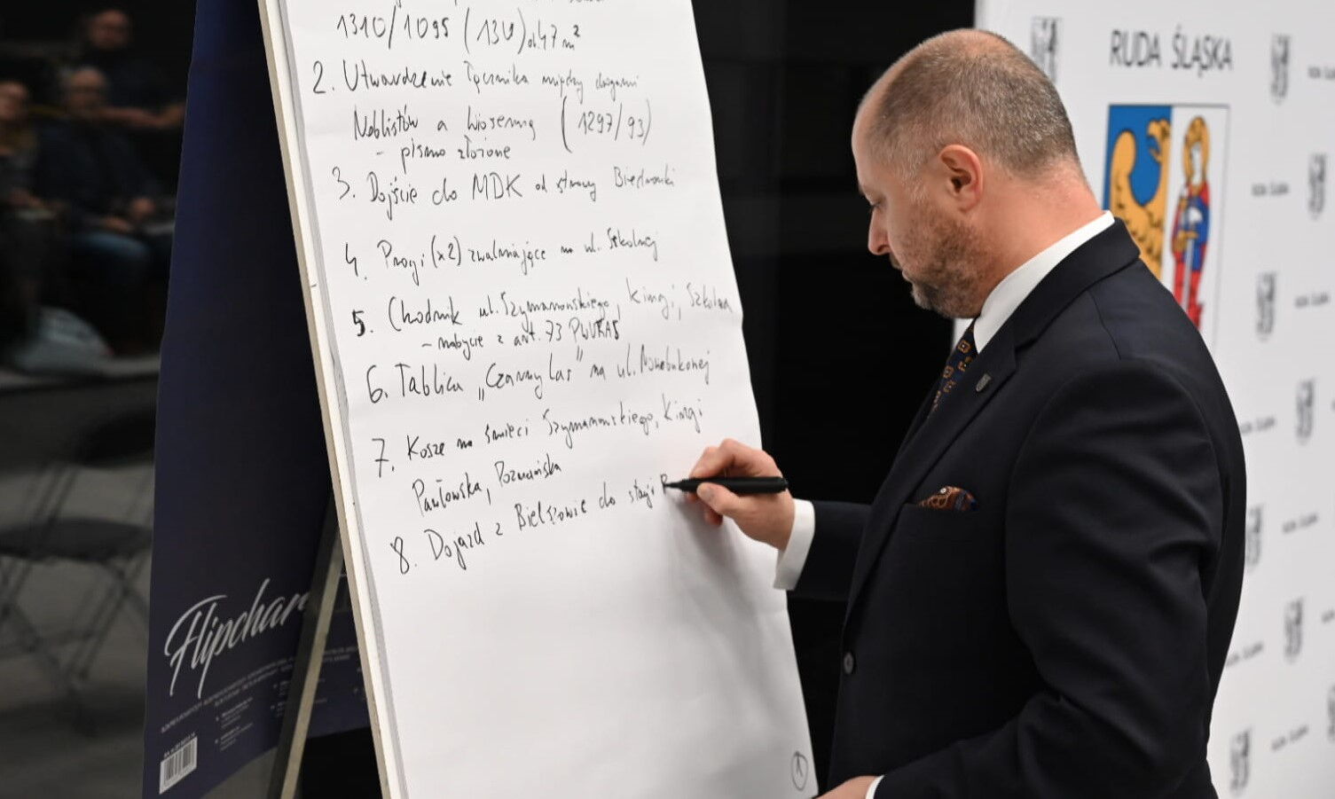 Mężczyzna w garniturze stoi przy flipcharcie i pisze czarnym markerem.