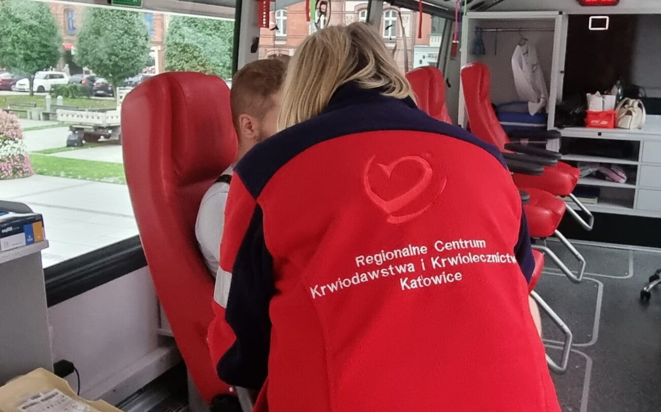 Kobieta stoi tyłem, na bluzie napis Regionalne centrum krwiodawstwa i krwiolecznictwa. Obok mężczyzna na fotelu oddaje krew.