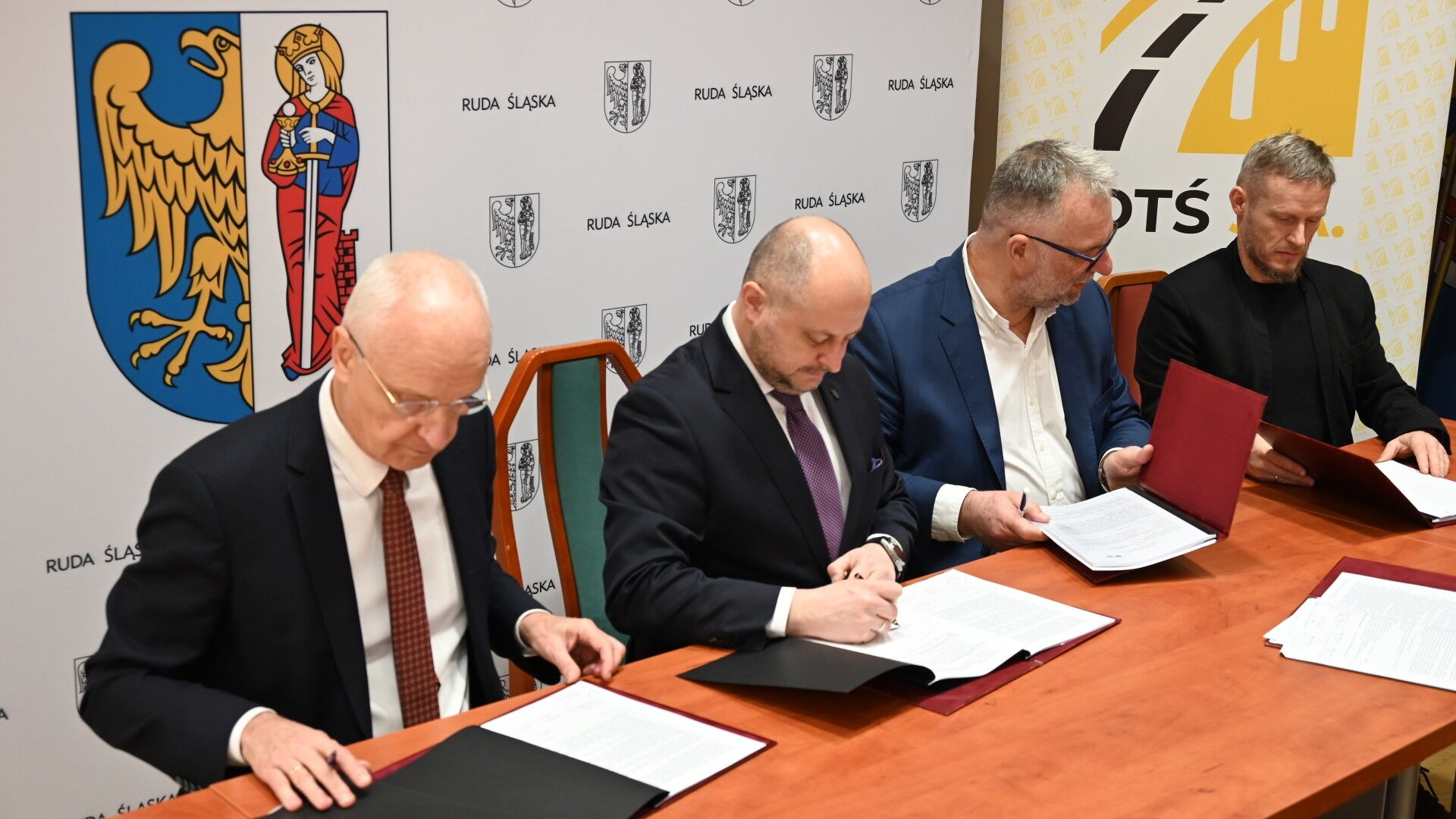Czterech mężczyzn podpisuje dokumenty. W tle ścianki z herbem Rudy Śląskiej logotyprem DTŚ S.A.