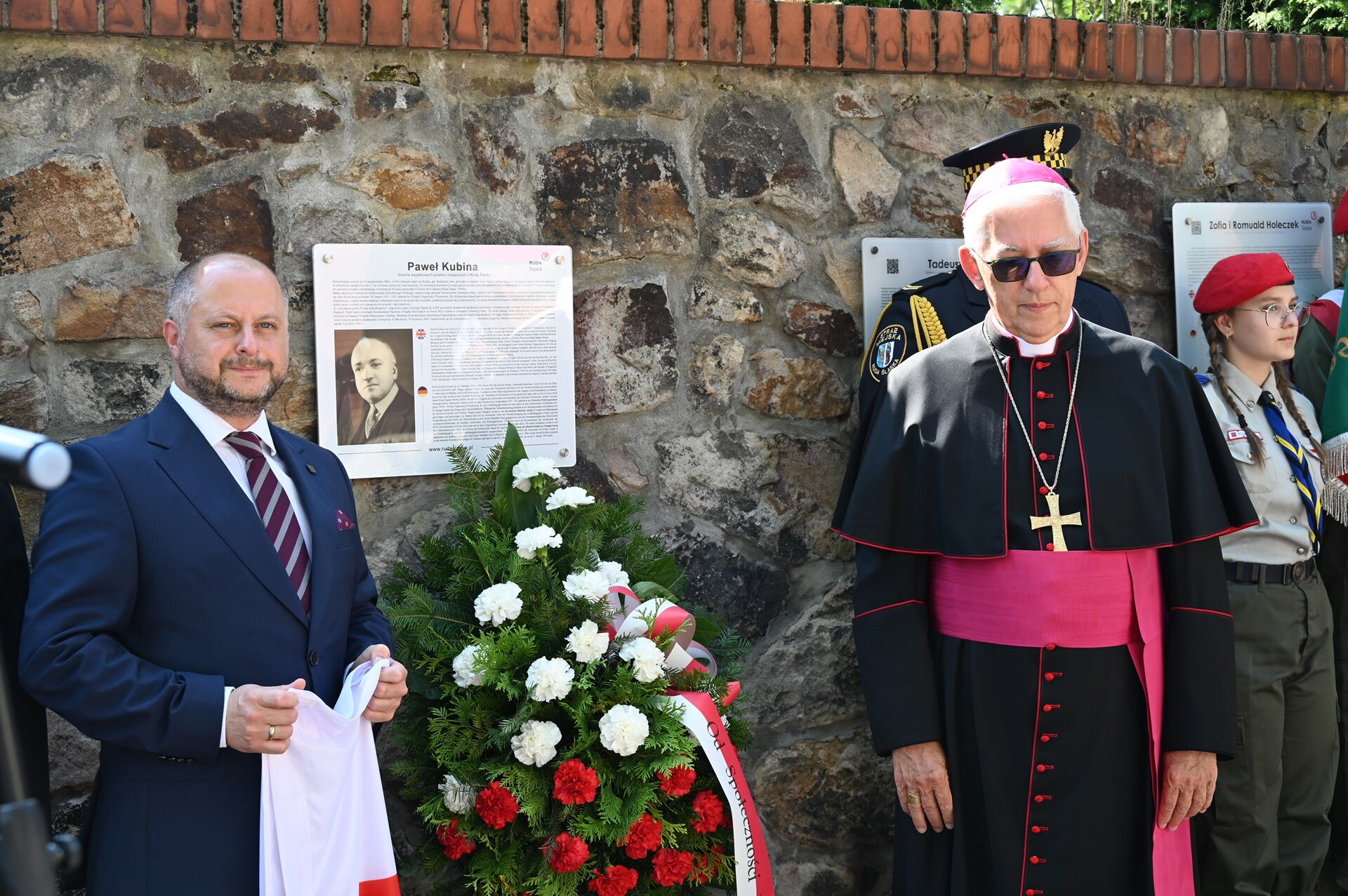 Na murze tablica, pod którą stoi wieniec z biało-czerwonych goździków, obok stoi mężczyzna w garniturze, z drugiej strony stoi arcybiskup oraz harcerka.