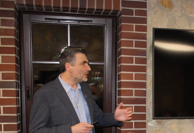 mężczyzna na tle okna zwrócony w prawą stronę, tłumaczy coś