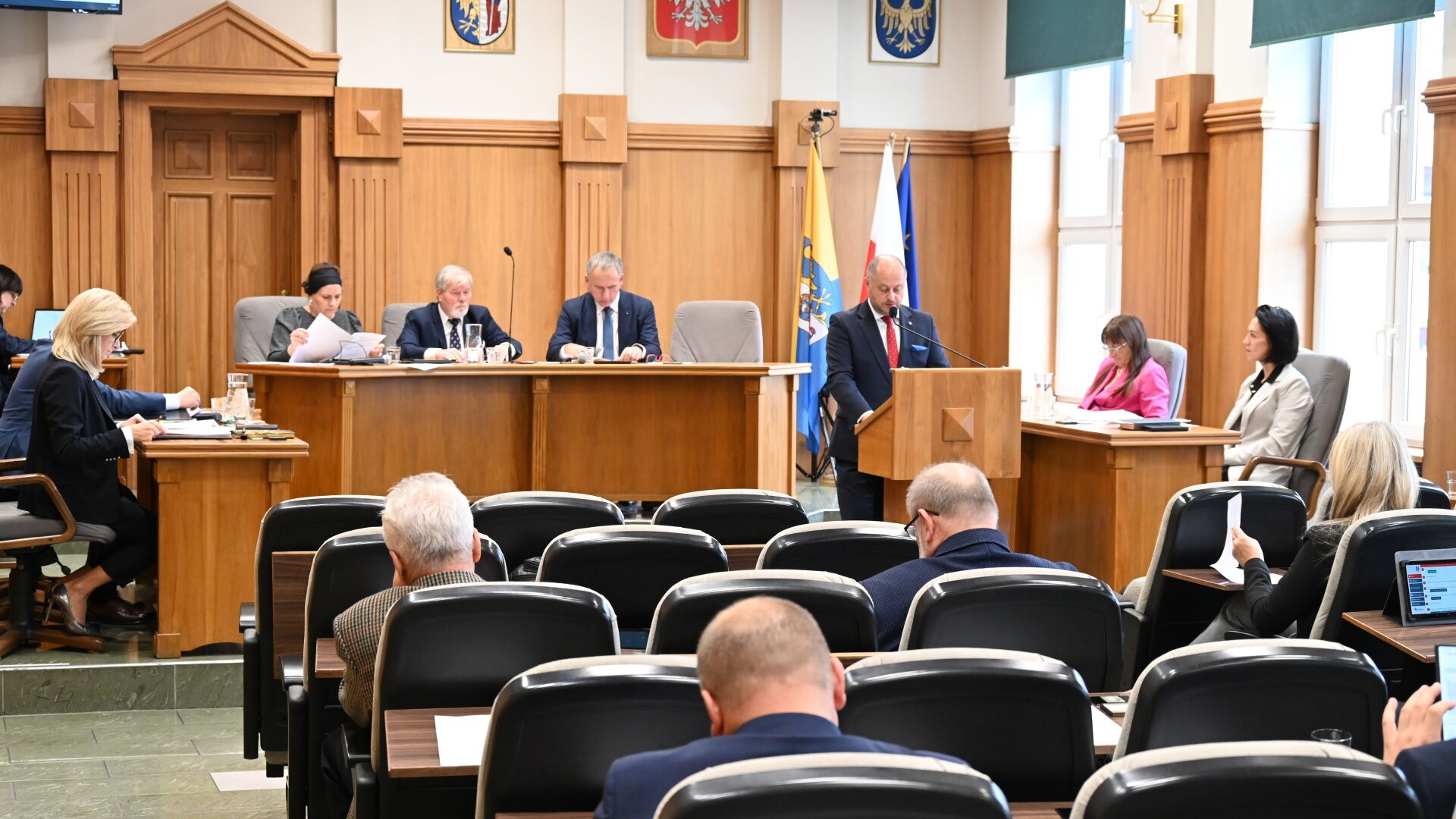 Sala sesyjna. Grupa osób siedzących na fotelach, w tle mężczyzna przy mównicy i kilka osób za stołem prezydialnym. Z tyłu flagi Unii Europejskiej, Polski, i Rudy Śląskiej.