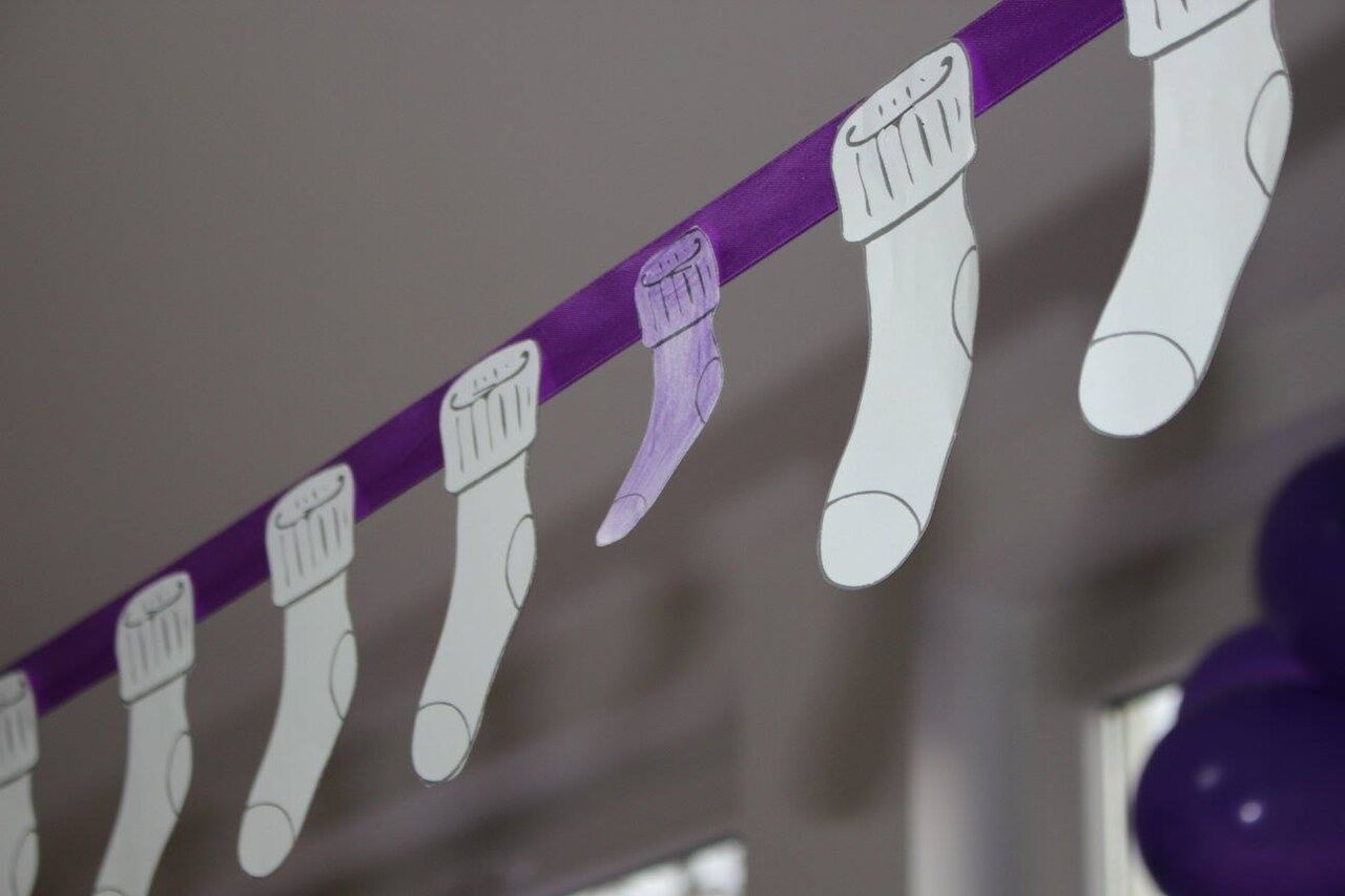 Do Fioletowej wstęgi przyczepione białe, papierowe skarpetki, jedna z nich jest mniejsza i fioletowa.