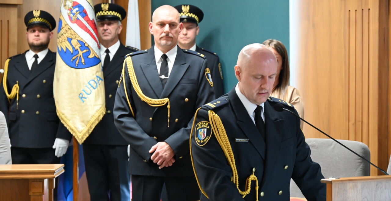 Strażnik miejski w mundueze przy mównicy, za nim kolejny strażnik w mundurze, w tle trzech strażników miejskich w mundurach ze sztandarem Rudy Śląskiej.