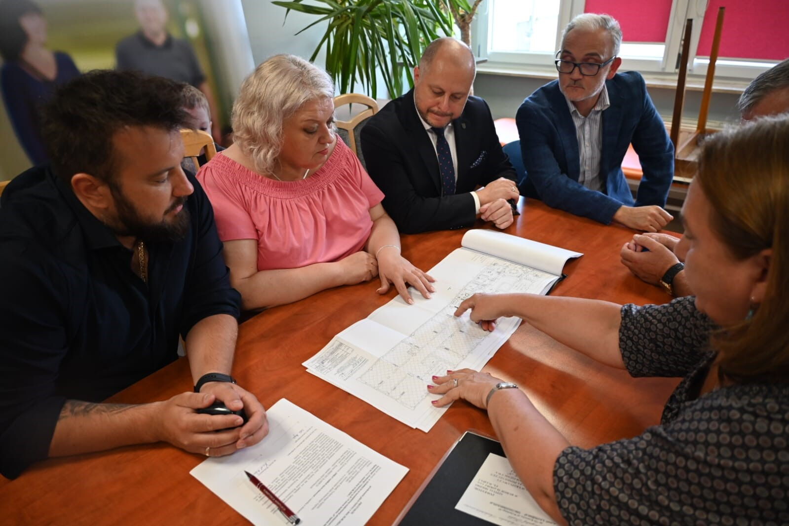 grupa osób siedzi przy stole nad rozłożonymi planami technicznymi