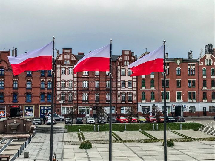 Na pierwszym planie 3 maszty z biało-czerwonymi flagami, na drugim planie zabytkowe kamienice.