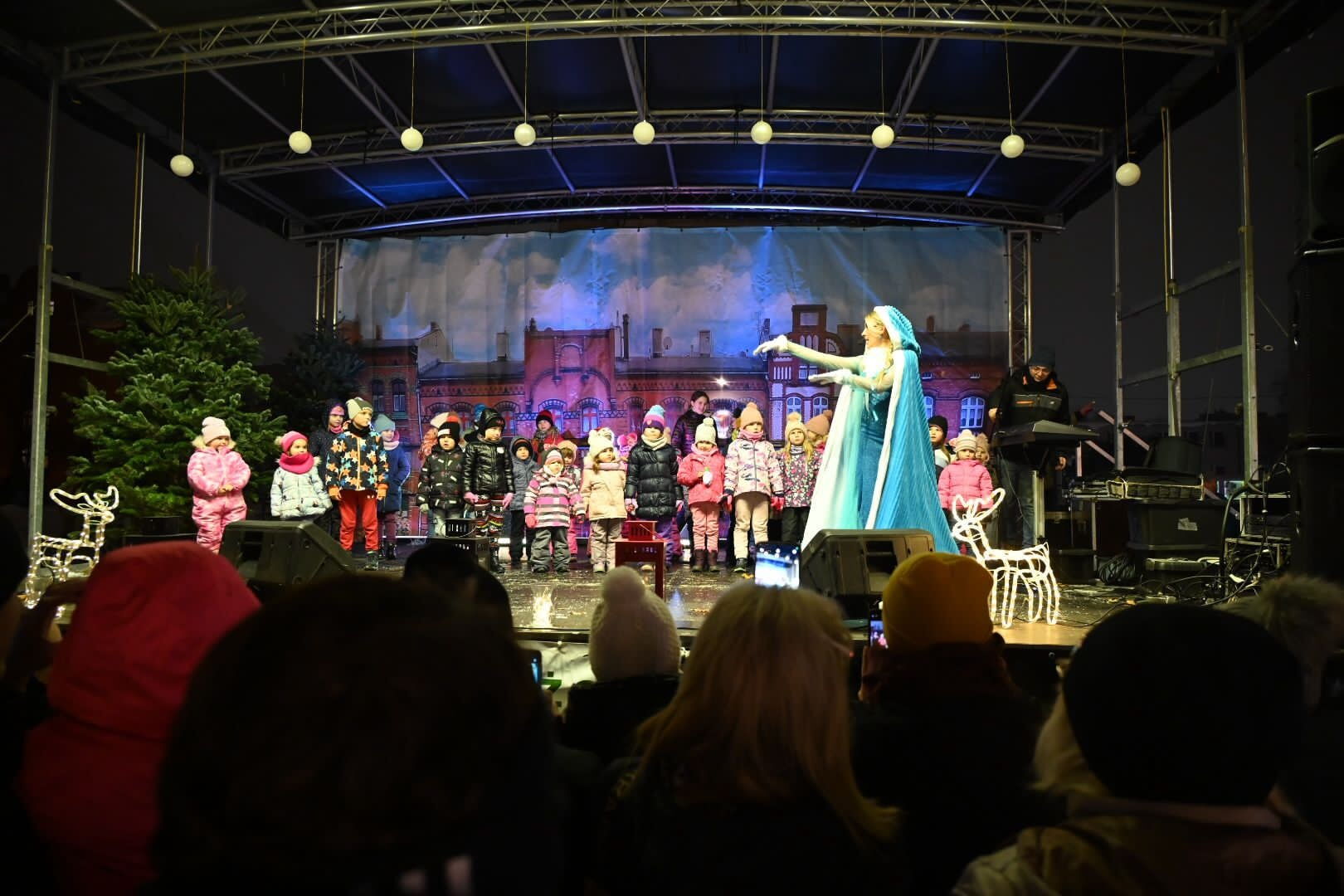 Na scenie duża grupa dzieci ubranych w zimowe ubrania oraz kobieta w bajkowym stroju Elzy.