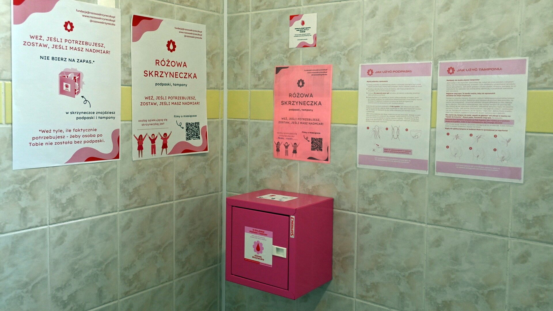 Różowa skrzynka na wykafelkowanej ścianie, nad nią materiały informacyjne akcji "Różowa Skrzyneczka".