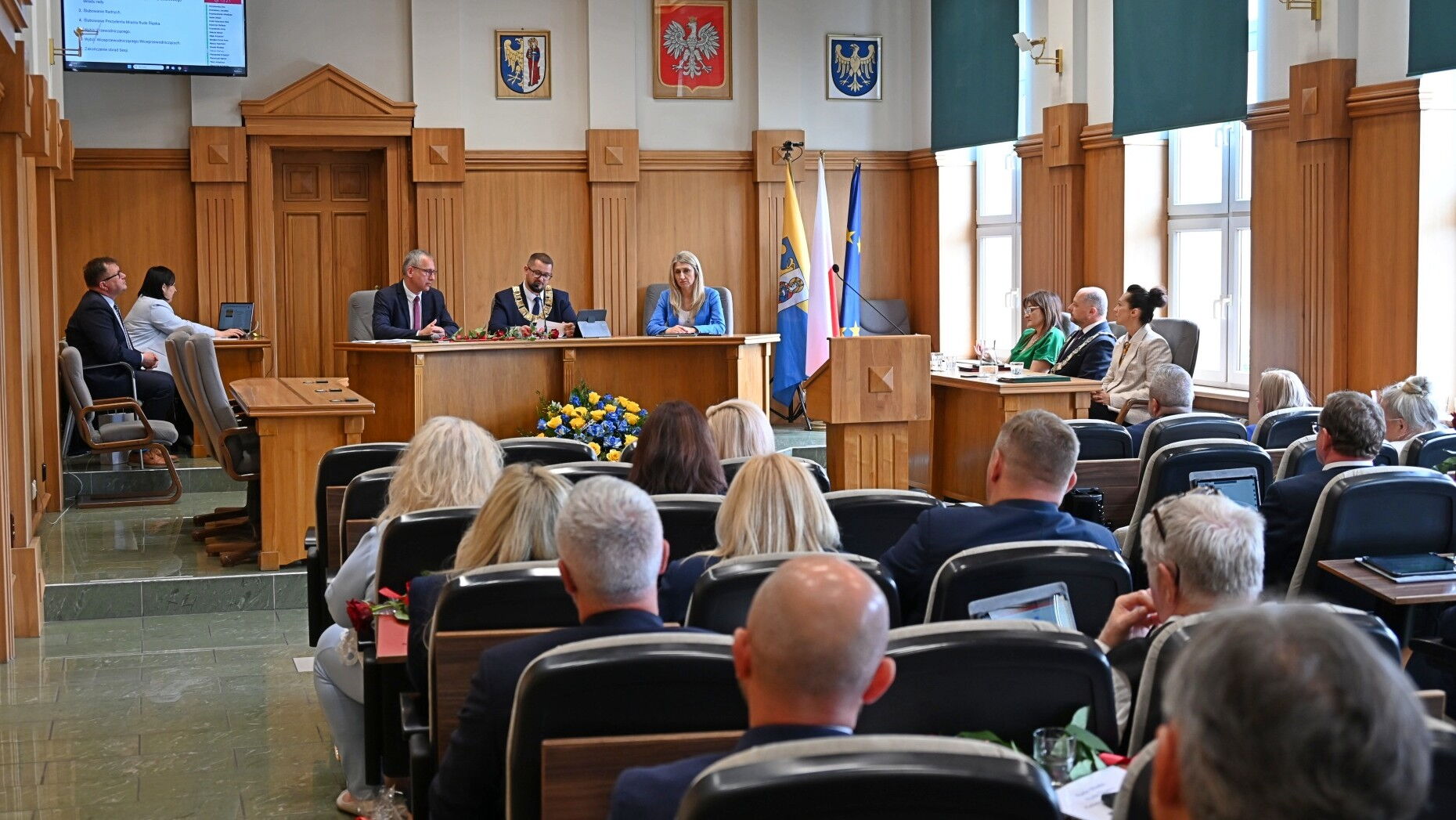 Sala obrad. Grupa osób siedzących na fotelach, mównica, kilka osób za stołem prezydialnym. Z tyłu flagi Unii Europejskiej, Polski, i Rudy Śląskiej oraz godła Rudy Śląskiej, Polski i Województwa Śląskiego..