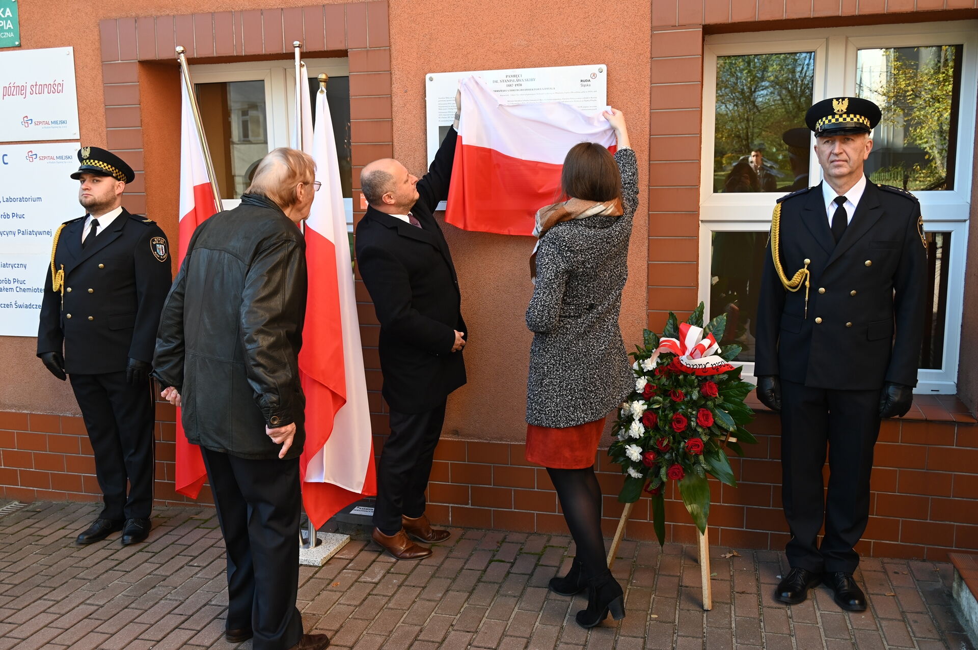 Mężczyzna i kobieta odsłaniają tablicę przymocowaną do ściany budynku. Obok stoi dwóch mundurowych oraz mężczyzna w cywilu. Widać wiązankę i biało-czerwone flagi.