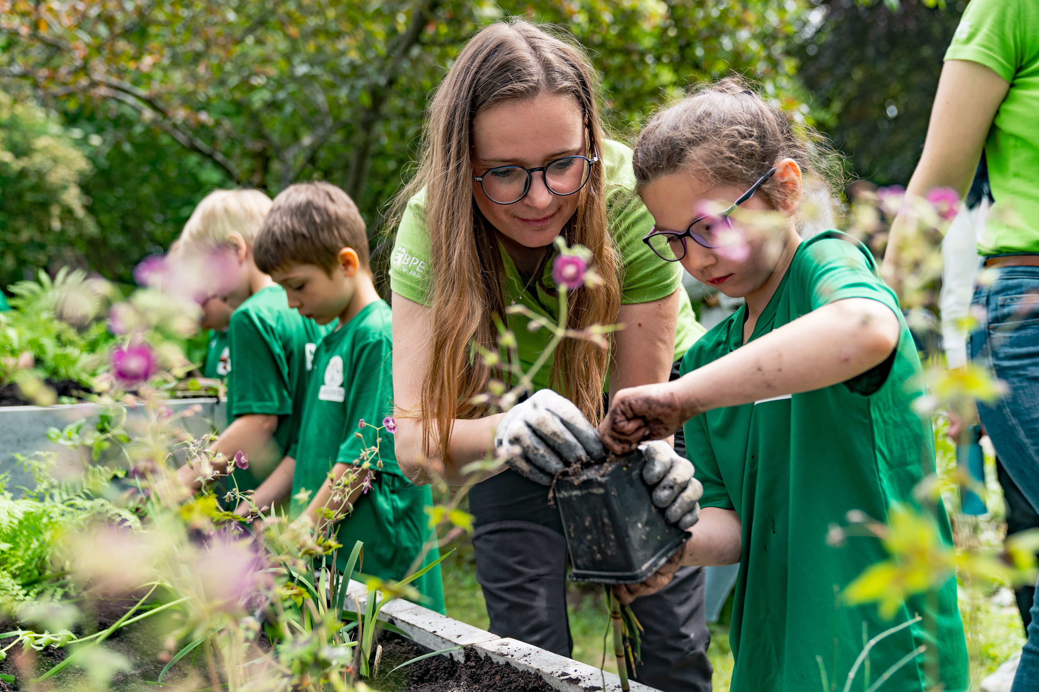 Sadzenie roślin - na pierwszym planie kobieta i dzieci z doniczką, w tle inne osoby. Z przodu rosnące kwiaty.