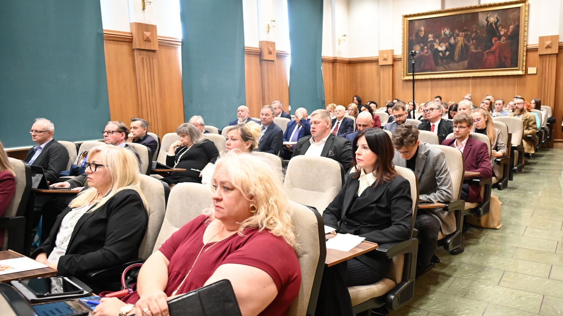 Grupa osób siedzi w fotelach na sali obrad. W tle ściana z obrazem.