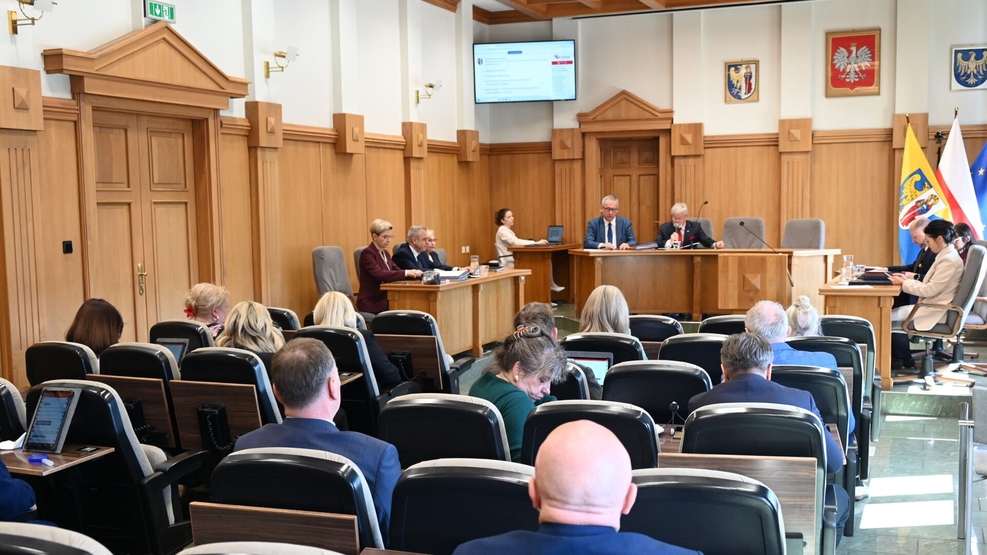 Sala obrad. Grupa osób siedzących na fotelach, mównica, kilka osób za stołem prezydialnym. Z tyłu flagi Unii Europejskiej, Polski, i Rudy Śląskiej oraz godła Rudy Śląskiej, Polski i Województwa Śląskiego.