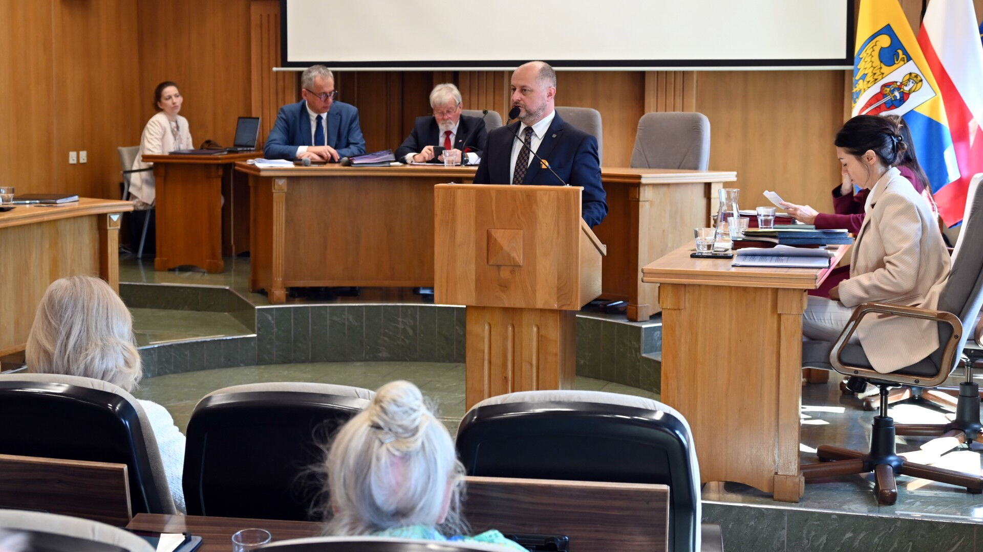 Sala obrad. Mężczyzna przy mównicy, kilka osób za stołem prezydialnym, z tyłu flagi Rudy Śląskiej i Polski.