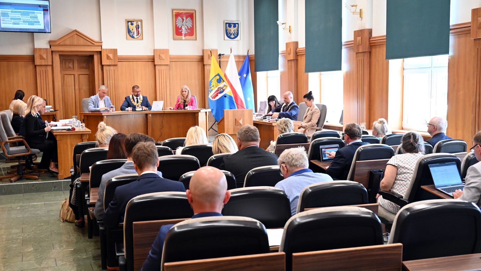 Sala obrad. Grupa osób siedzących na fotelach, mównica, kilka osób za stołem prezydialnym. Z tyłu flagi Unii Europejskiej, Polski, i Rudy Śląskiej oraz godła Rudy Śląskiej, Polski i Województwa Śląskiego..