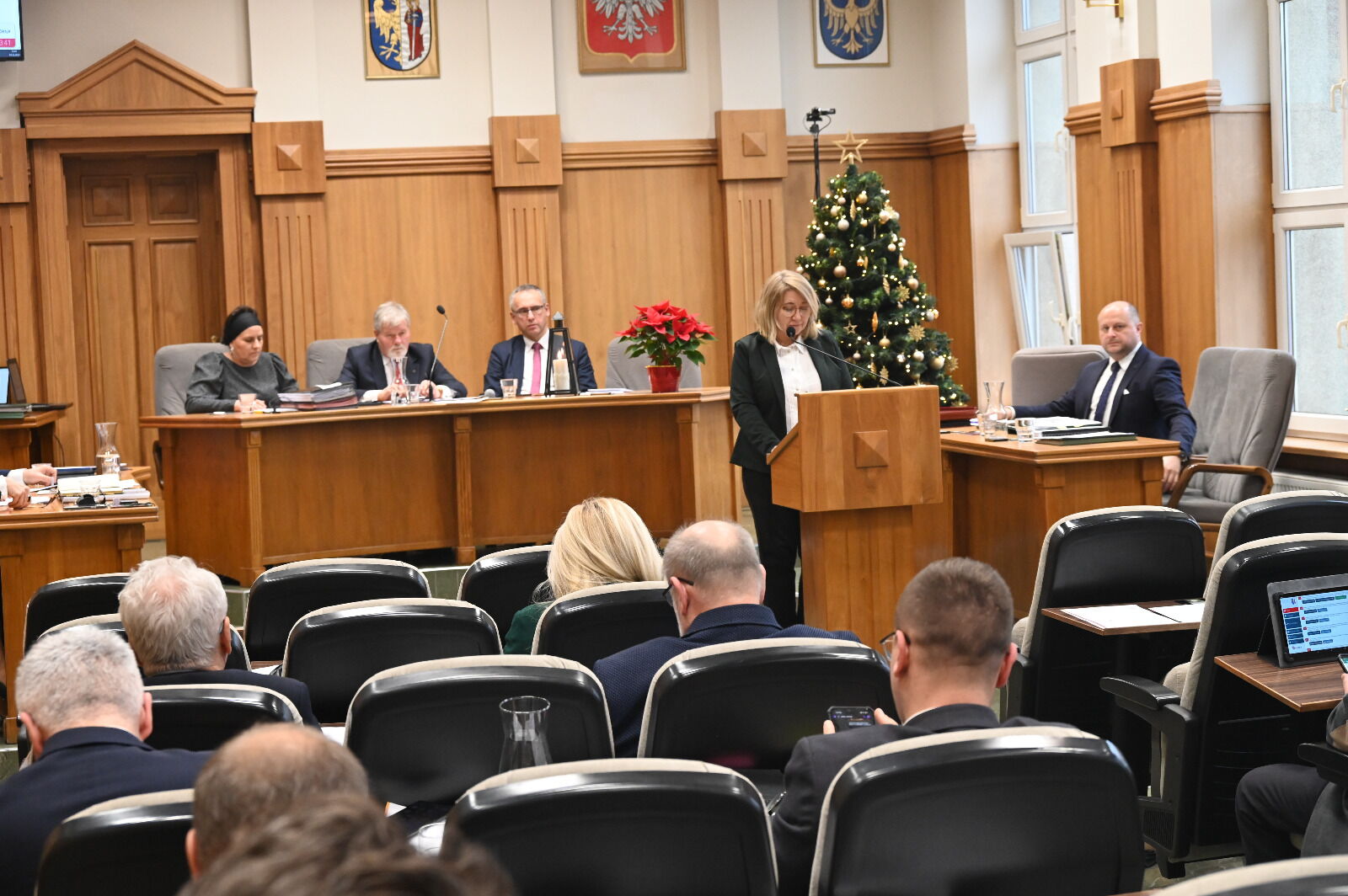 Sala sesyjna. Kilka osób widzianych z tyłu siedzi na krzesłach. Kobieta stoi przy mównicy i mikrofonie. Za stołem prezydialnym siedzą 3 osoby. Po prawej stronie choinka.