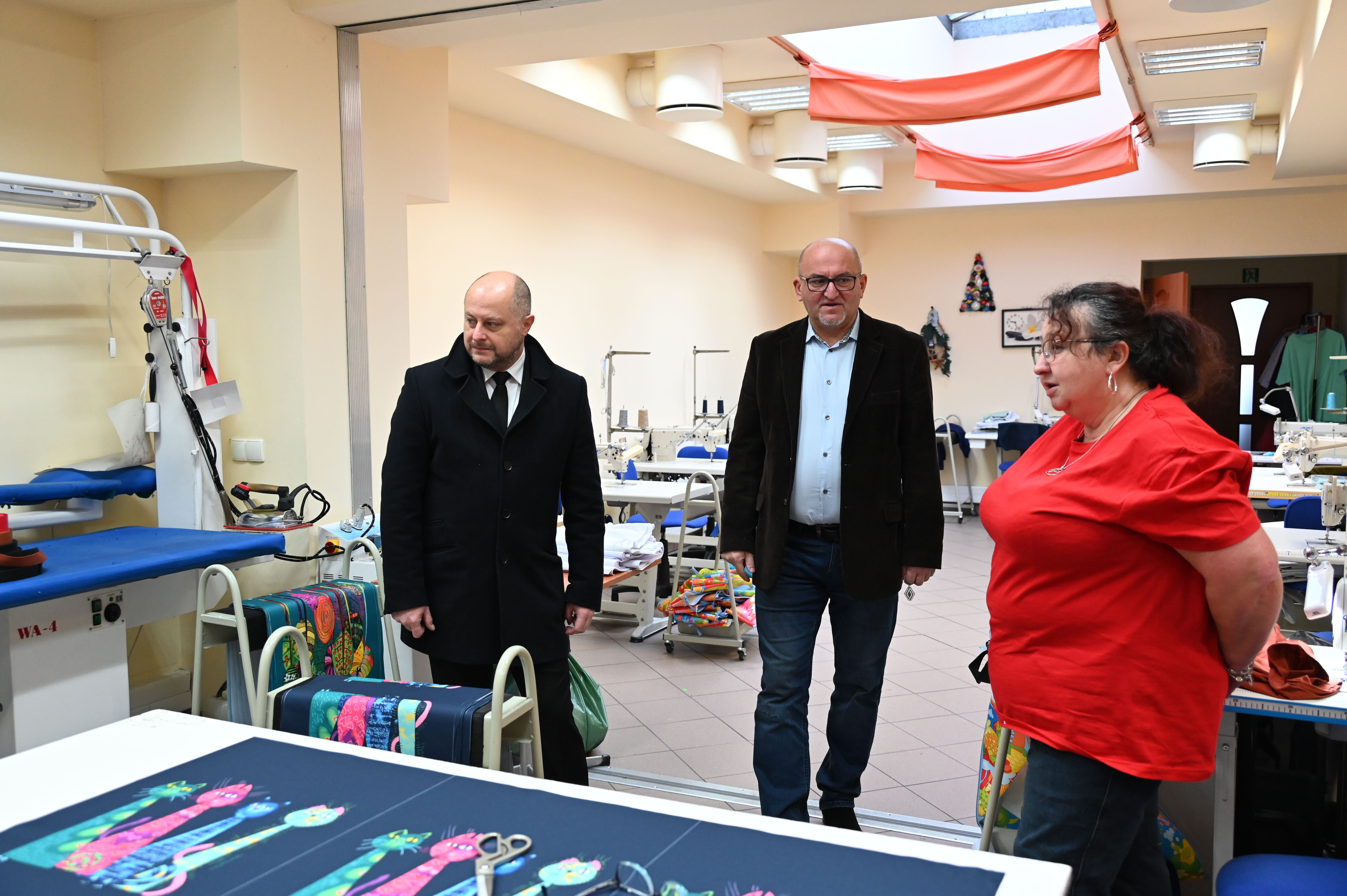 w pomieszczeniu trzy osoby przyglądają się maszynom i procesowi produkcji w szwalni, na stole kolorowe tkaniny, w tle materiały i urządzenia