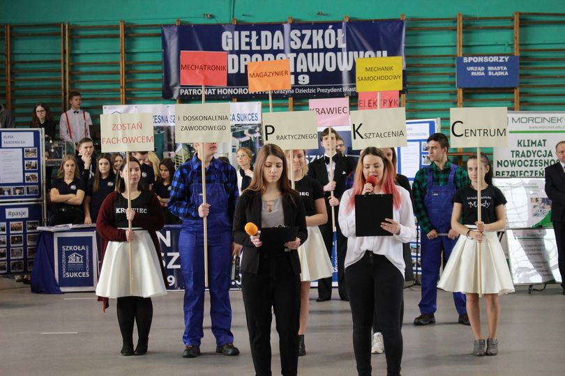 Grupa osób stoi na terenie hali sportowej. Z przodu dwie dziewczyny z mikrofonami. Osoby z tyłu trzymają plakaty.