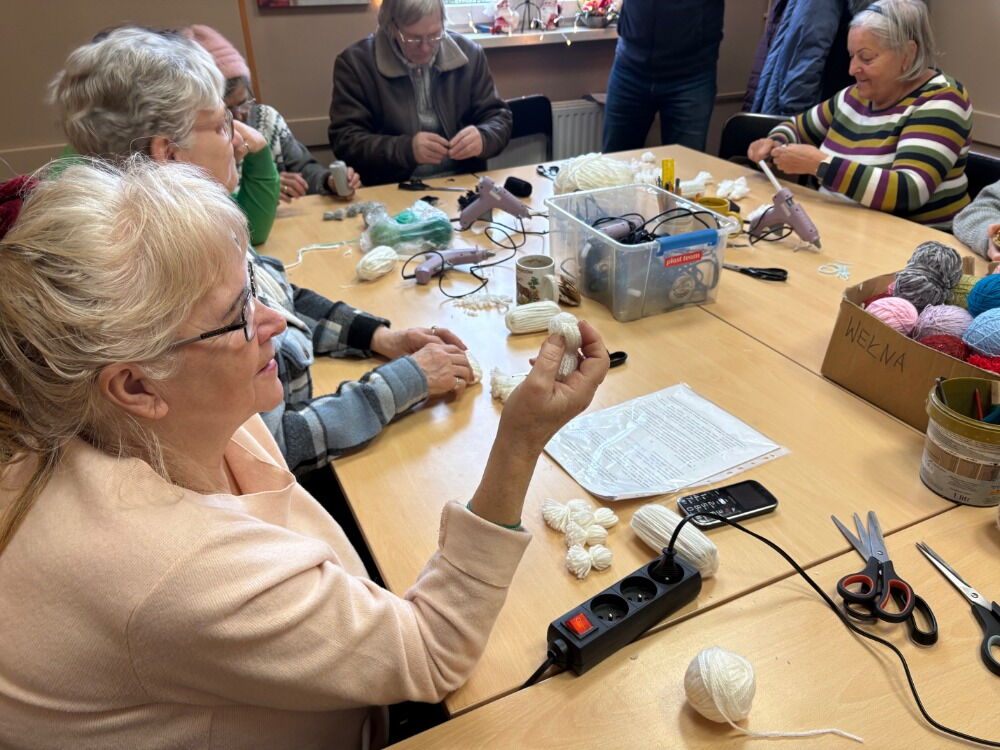 Na stole wiele materiałów do rękodzieła. Obok stołu siedzą kobiety.