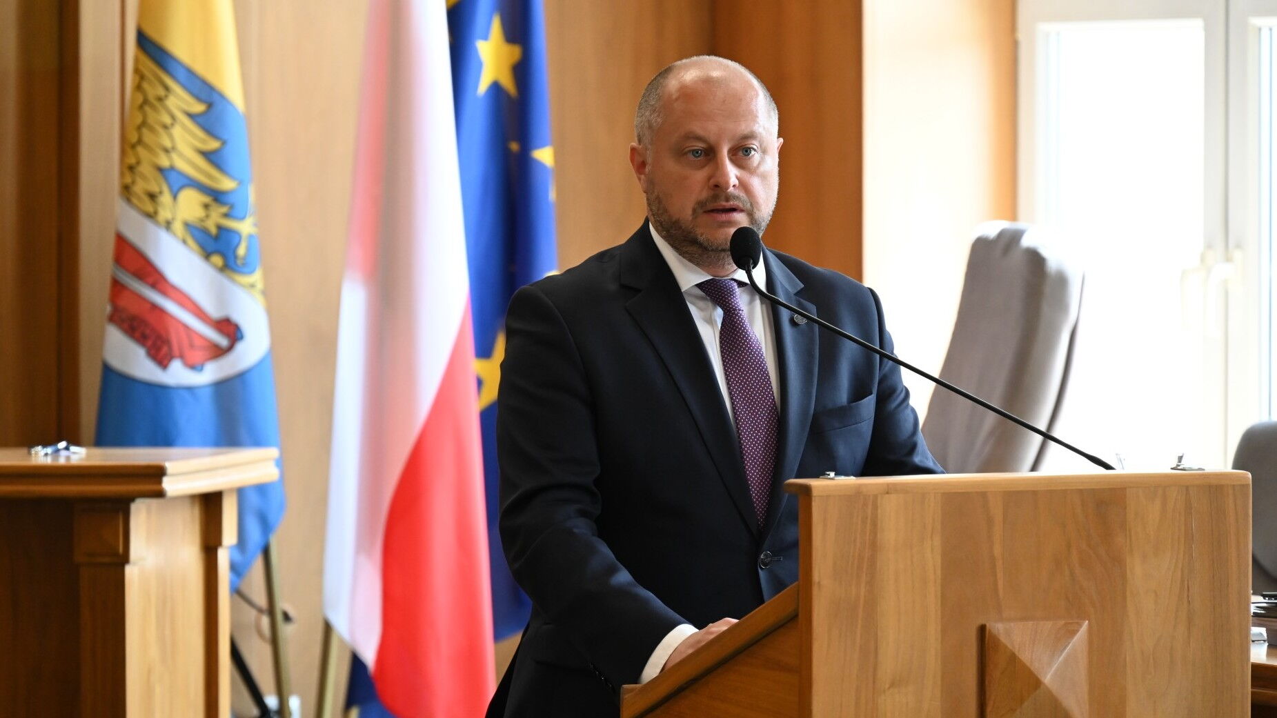 Mężczyzna w garniturze przy mównicy mówi do mikrofonu, w tle flagi Rudy Śląskiej, Polski i Unii Europejskiej.