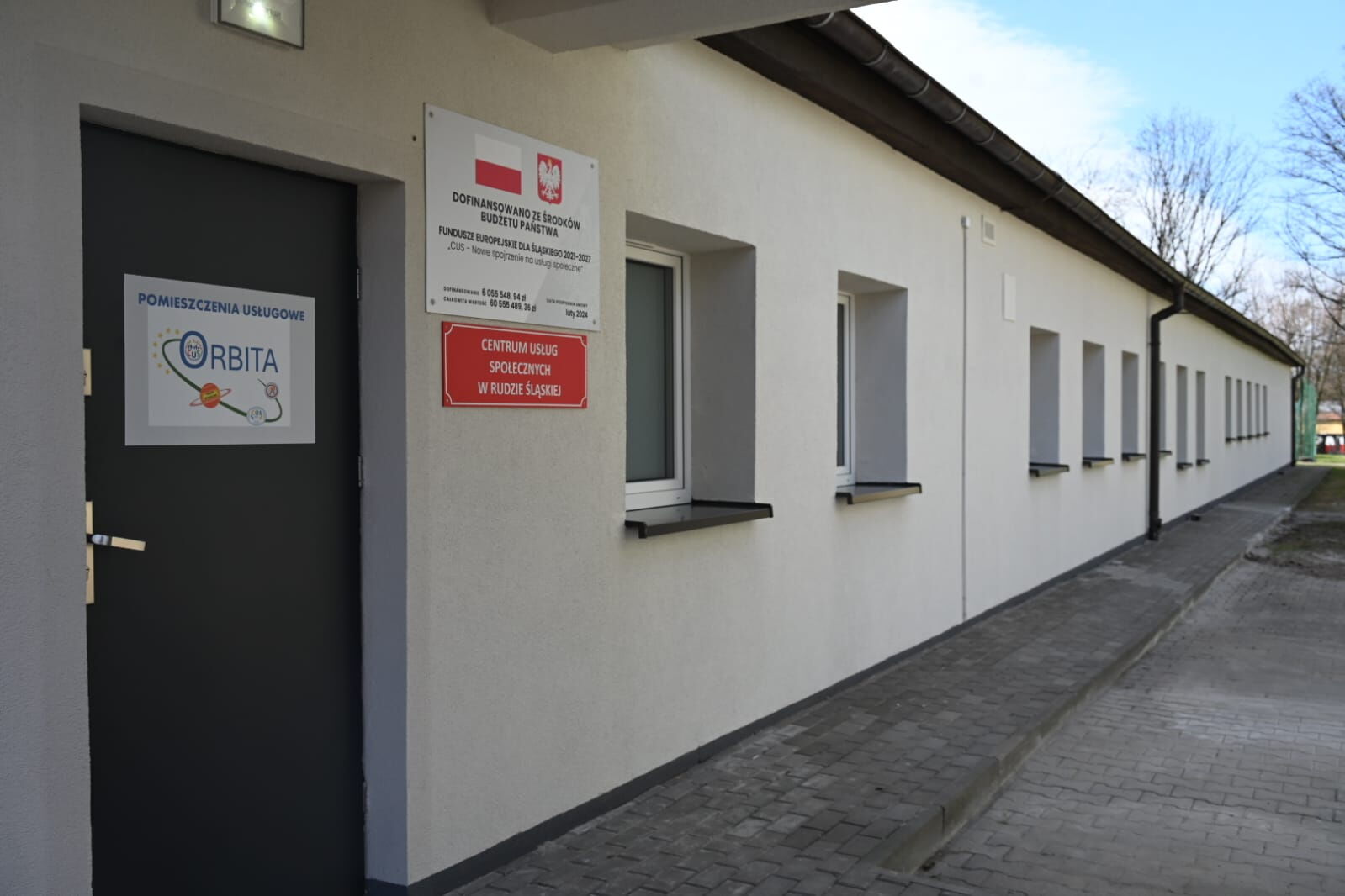 Parterowy jasny budynek, na którym zawieszone są tablice. W oddali widać drzewa.