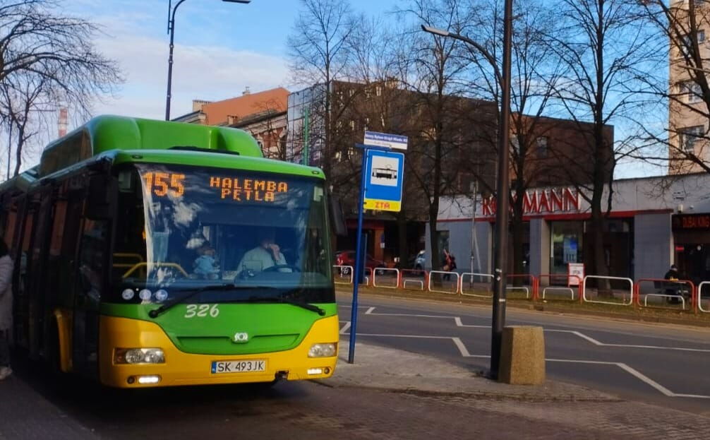 Autobus miejski linii 155. Obok znak drogowy, kosz, droga, drzewa. W tle budynki handlowe, bloki.