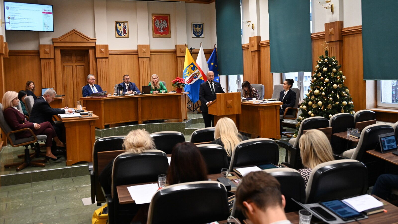 Sala sesyjna. Grupa osób siedzących na fotelach, w tle mężczyzna przy mównicy i kilka osób za stołem prezydialnym. Z tyłu flagi Unii Europejskiej, Polski, i Rudy Śląskiej, z boku choinka.