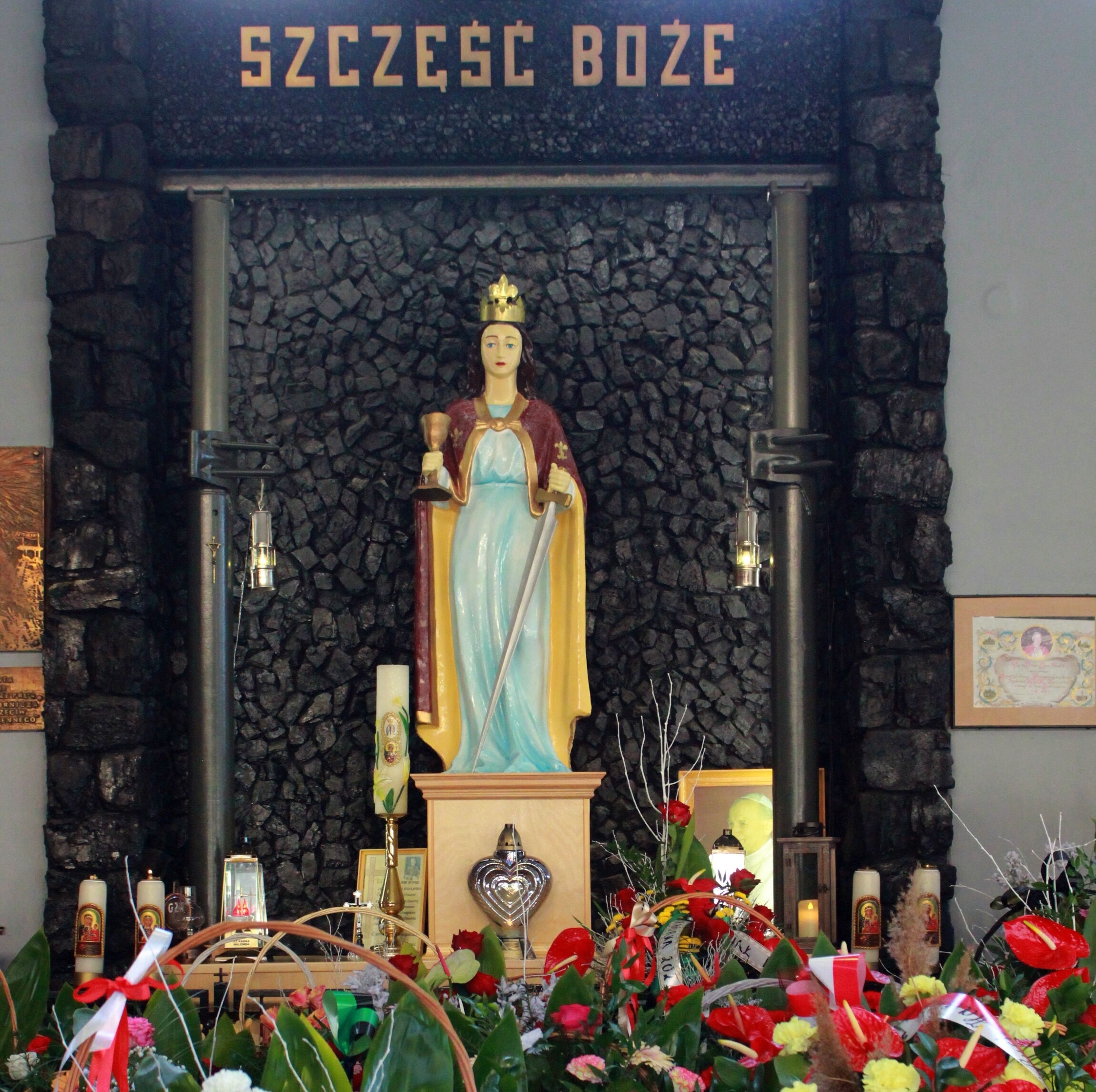 Statua przedstawiająca wizerunek świętej Barbary z kielichem i mieczem stojącej na tle ściany z węgla. Przed posągiem złożone są kwiaty i znicze.