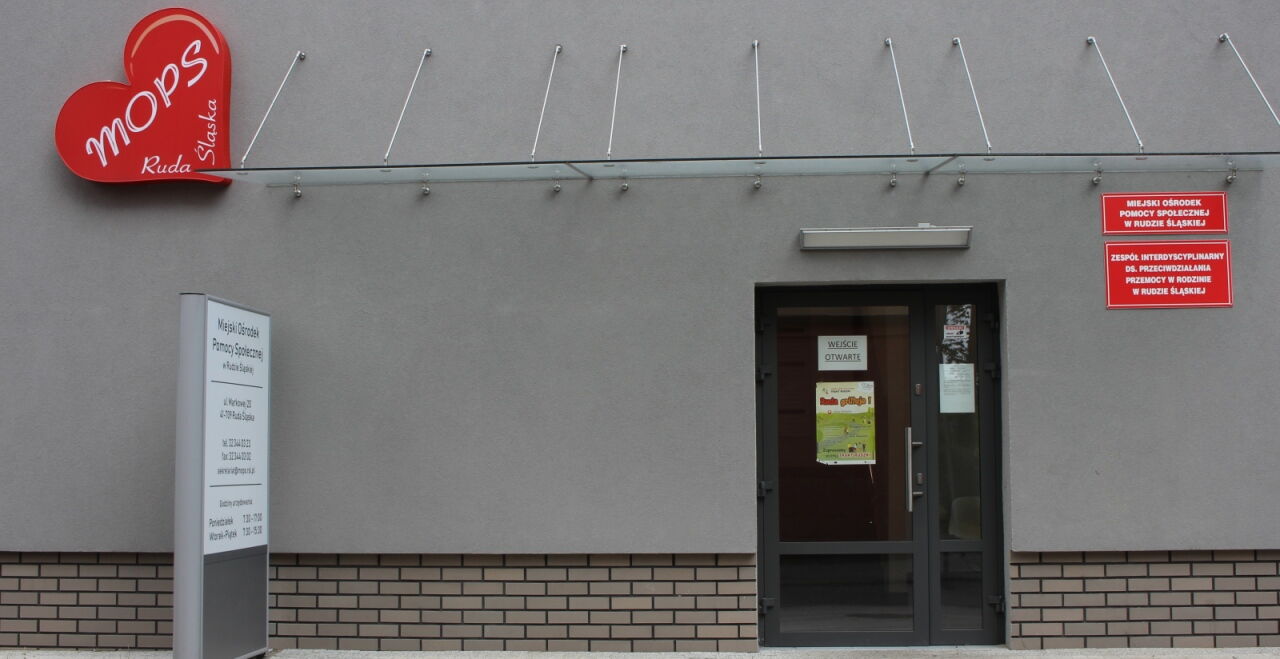 Wejście do szarego budynku. Na ścianie czerwone serce z białym napisem "MOPS Ruda Śląska" oraz czerwone tablice z białymi napisami. Przed budynkiem tablica informacyjna.