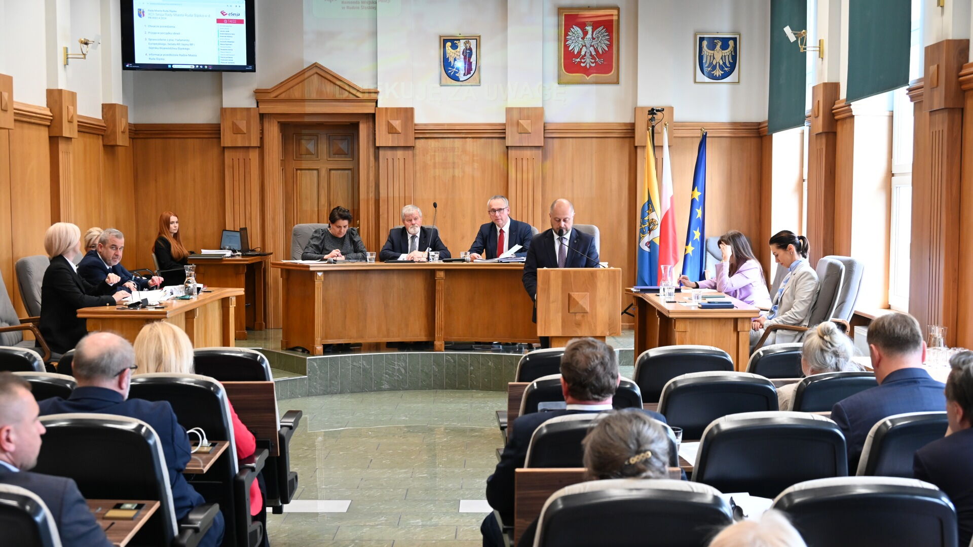 Sala sesyjna. Grupa osób siedzących na fotelach, w tle mężczyzna przy mównicy i kilka osób za stołem prezydialnym. Z tyłu flagi Unii Europejskiej, Polski, i Rudy Śląskiej.