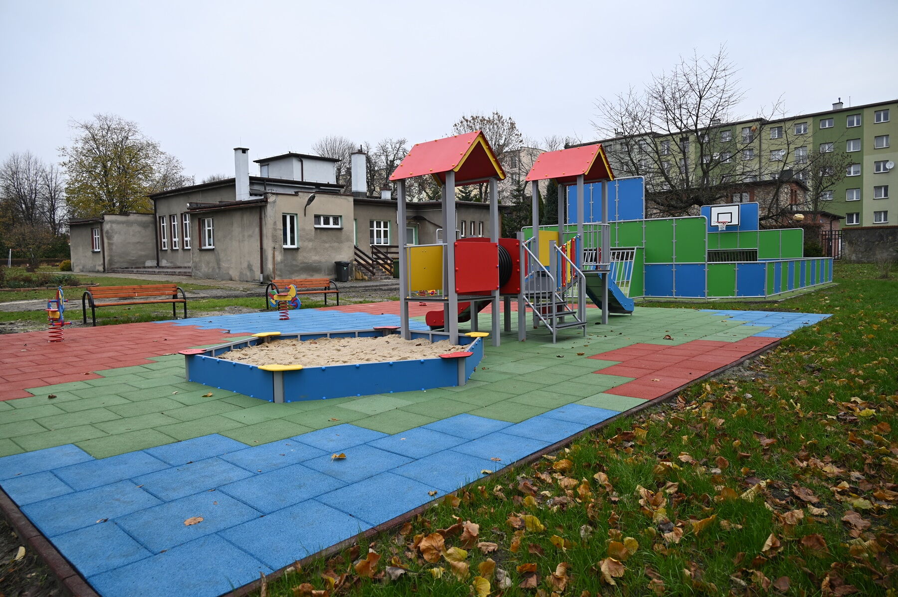 Na pierwszym planie kolorowy plac zabaw z piaskownicą. W tle szary, parterowy budynek.