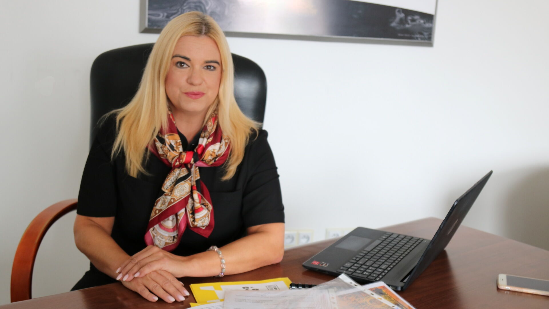 Kobieta siedzi na fotelu za biurkiem. Na biurku laptop, smartfon i dokumenty.