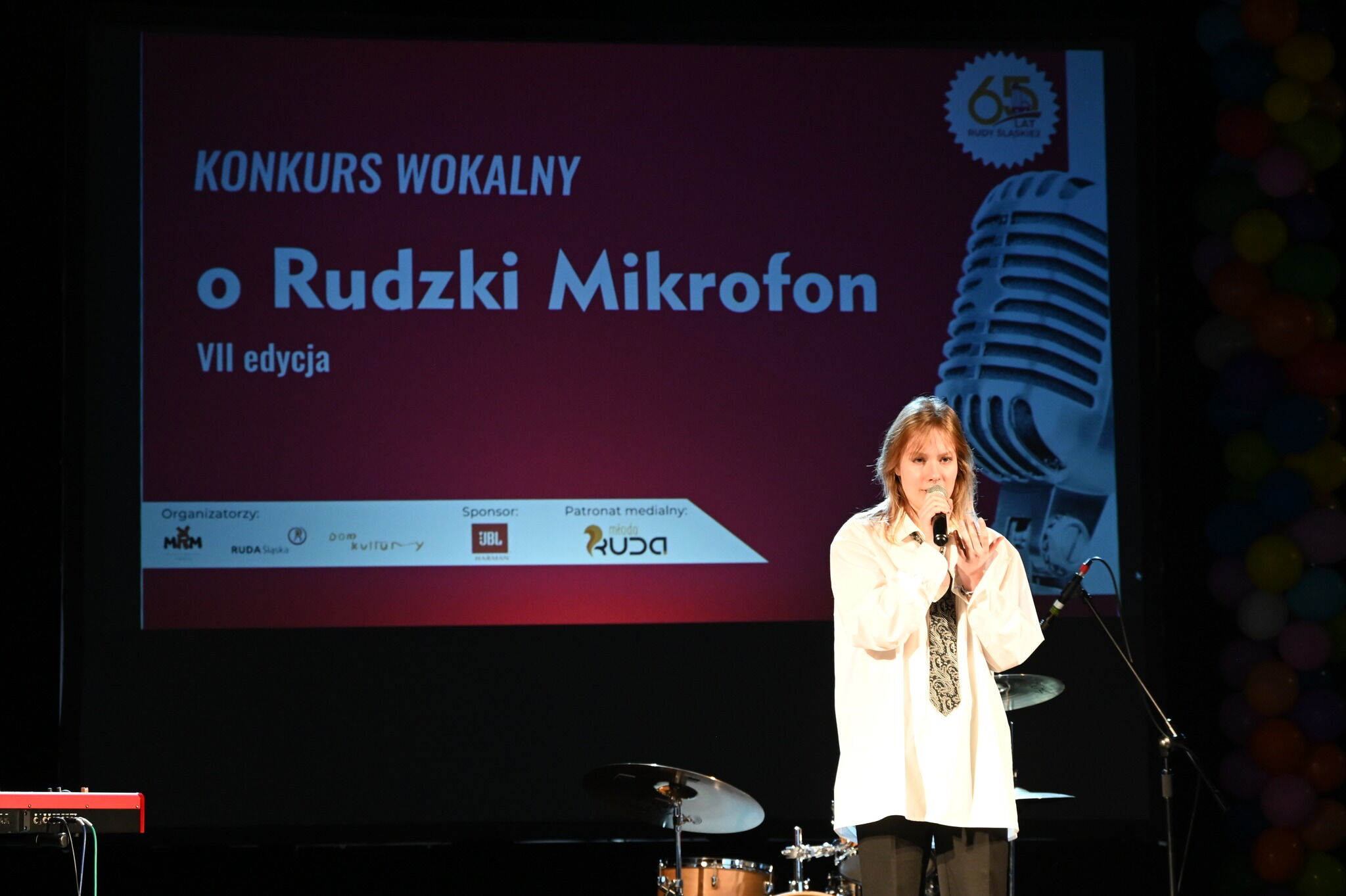 na scenie w pomieszczeniu młoda kobieta śpiewa do mikrofonu, w tle baner z napisem: konkurs wokalny o rudzki mikrofon