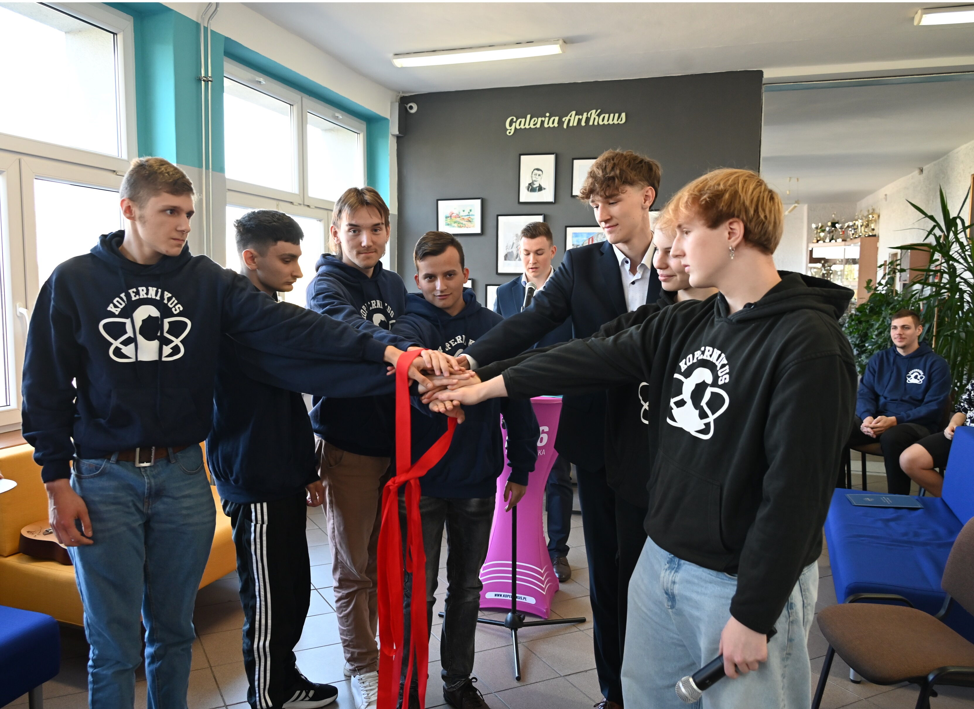 w pomieszczeniu grupa młodych osób stoi w kręgu i trzyma się za ręce. Ręce są obwiązane czerwoną wstążką