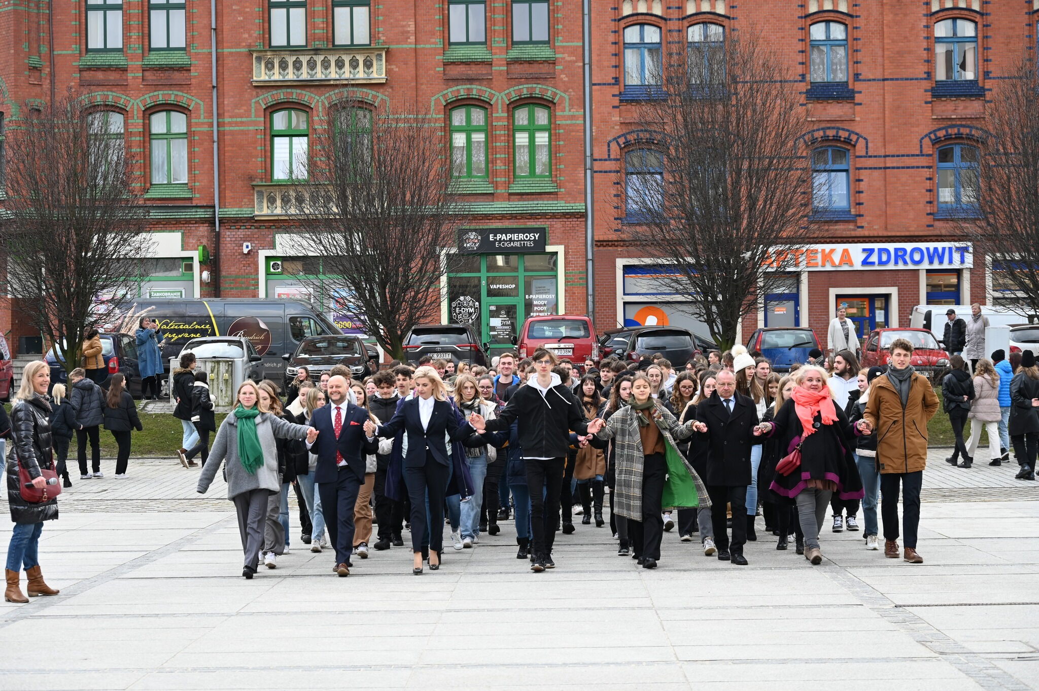 w plenerze, na rynku młodzież  tańczy poloneza w pierwszym rzędzie 4 pary trzymają się za ręce