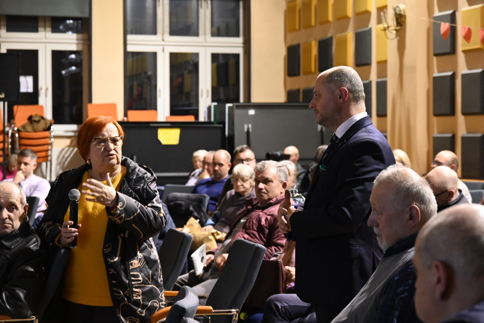 Kobieta z mikrofonem, obok słuchający jej mężczyzna w garniturze, wokół grupa ludzi siedzących na krzesłach.