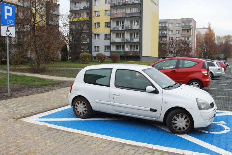Samochód stojący na niebieskim polu parkingowym dla osób z niepełnosprawnością. Obok znak miejsca parkingowego dla osób z niepełnosprawnością. W tle samochody i budynki.