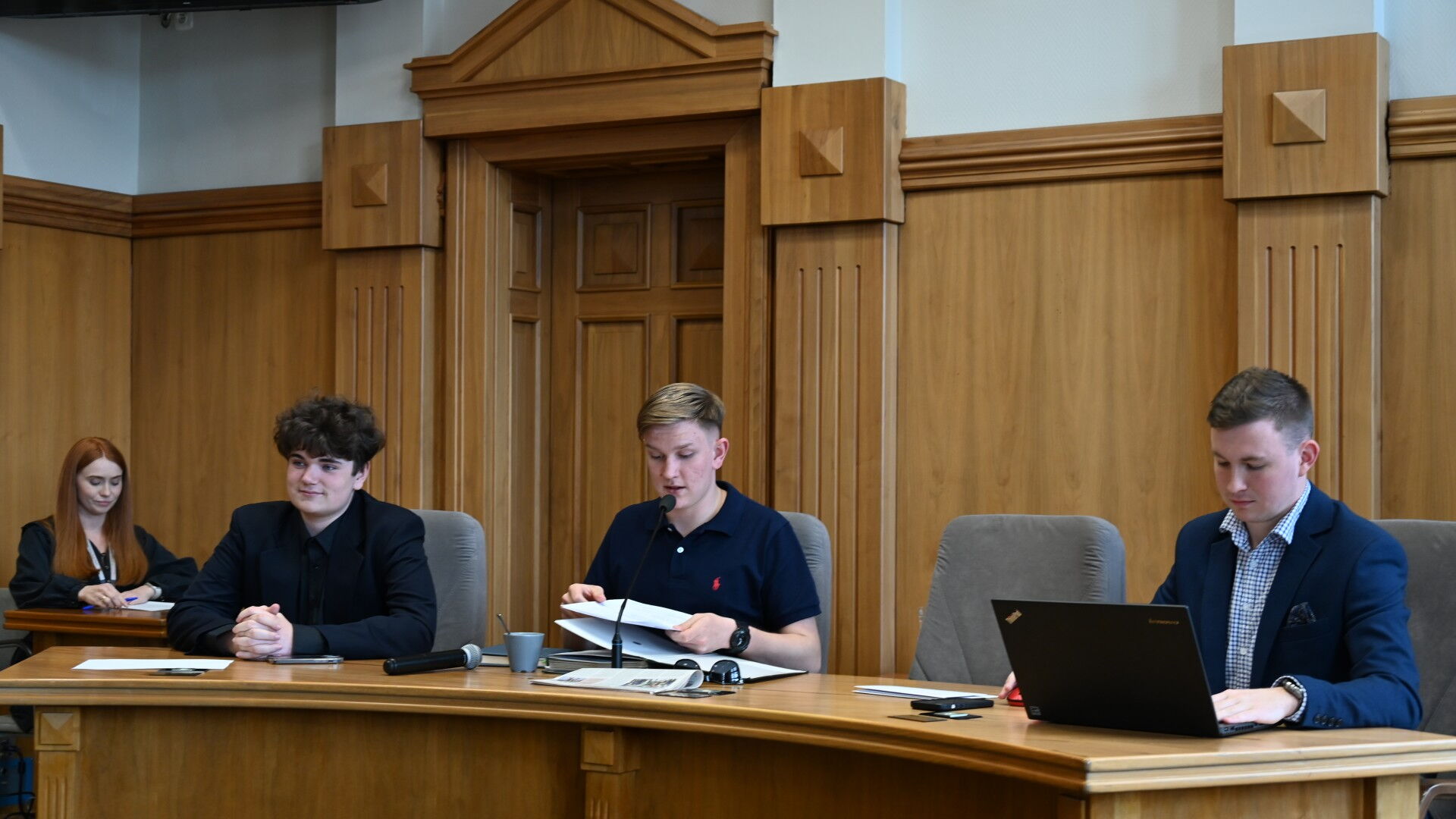 Trzech mężczyzn za stołem prezydialnym, w tle kobieta przy stoliku.