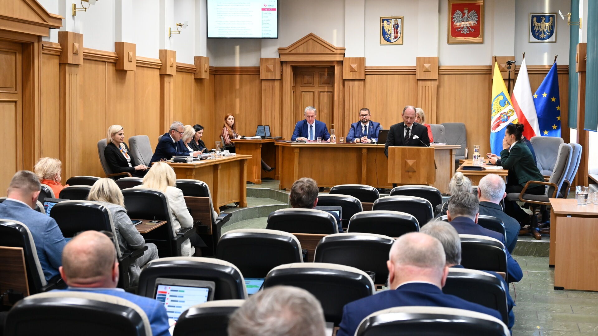 Sala sesyjna. Grupa osób siedzących na fotelach, w tle mężczyzna przy mównicy i kilka osób za stołem prezydialnym. Z tyłu flagi Unii Europejskiej, Polski, i Rudy Śląskiej.