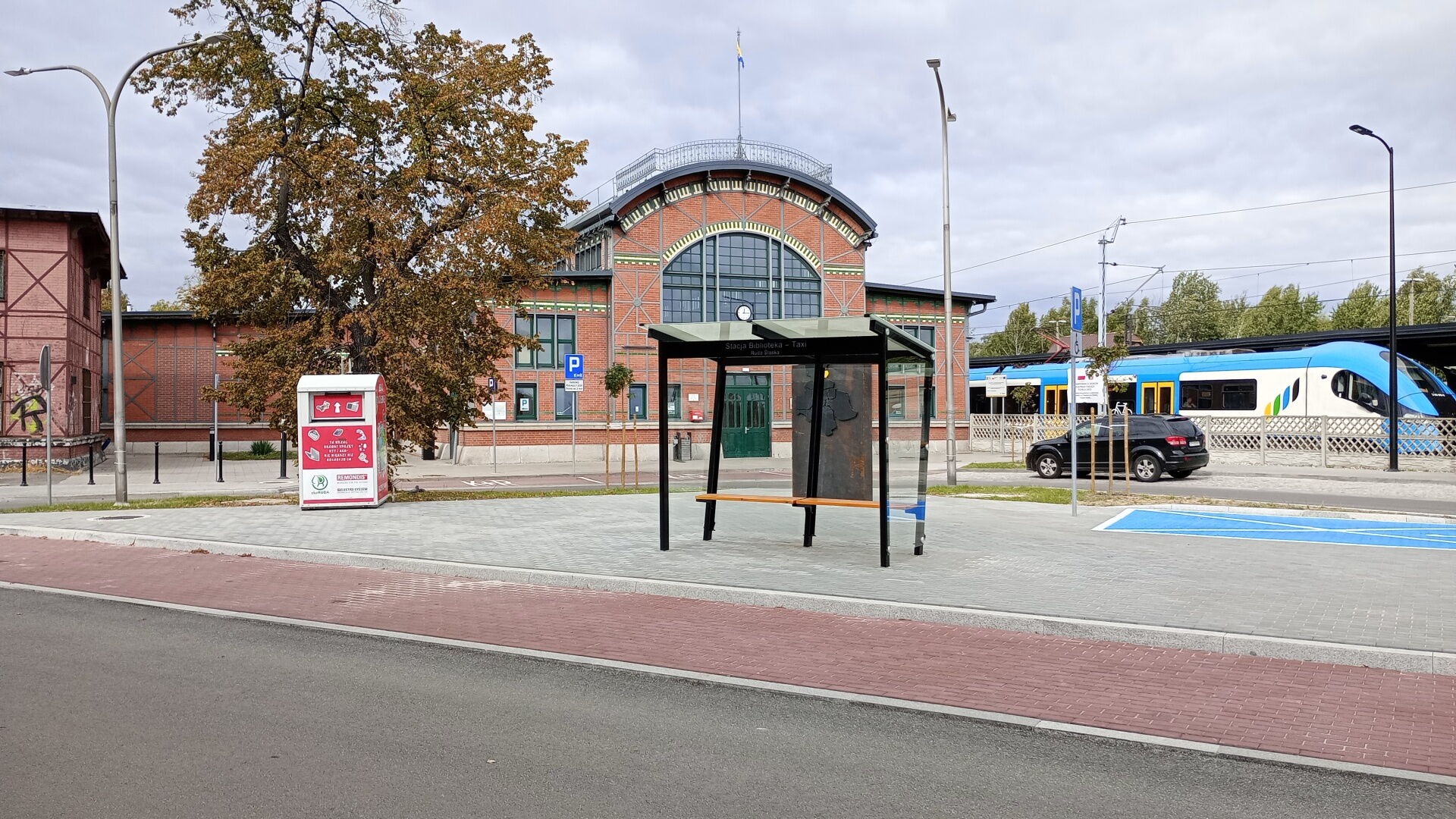 na pierwszym planie nowa droga , ścieżka rowerowa i przystanek autobusowy. W tle budynek dworca i pociąg.