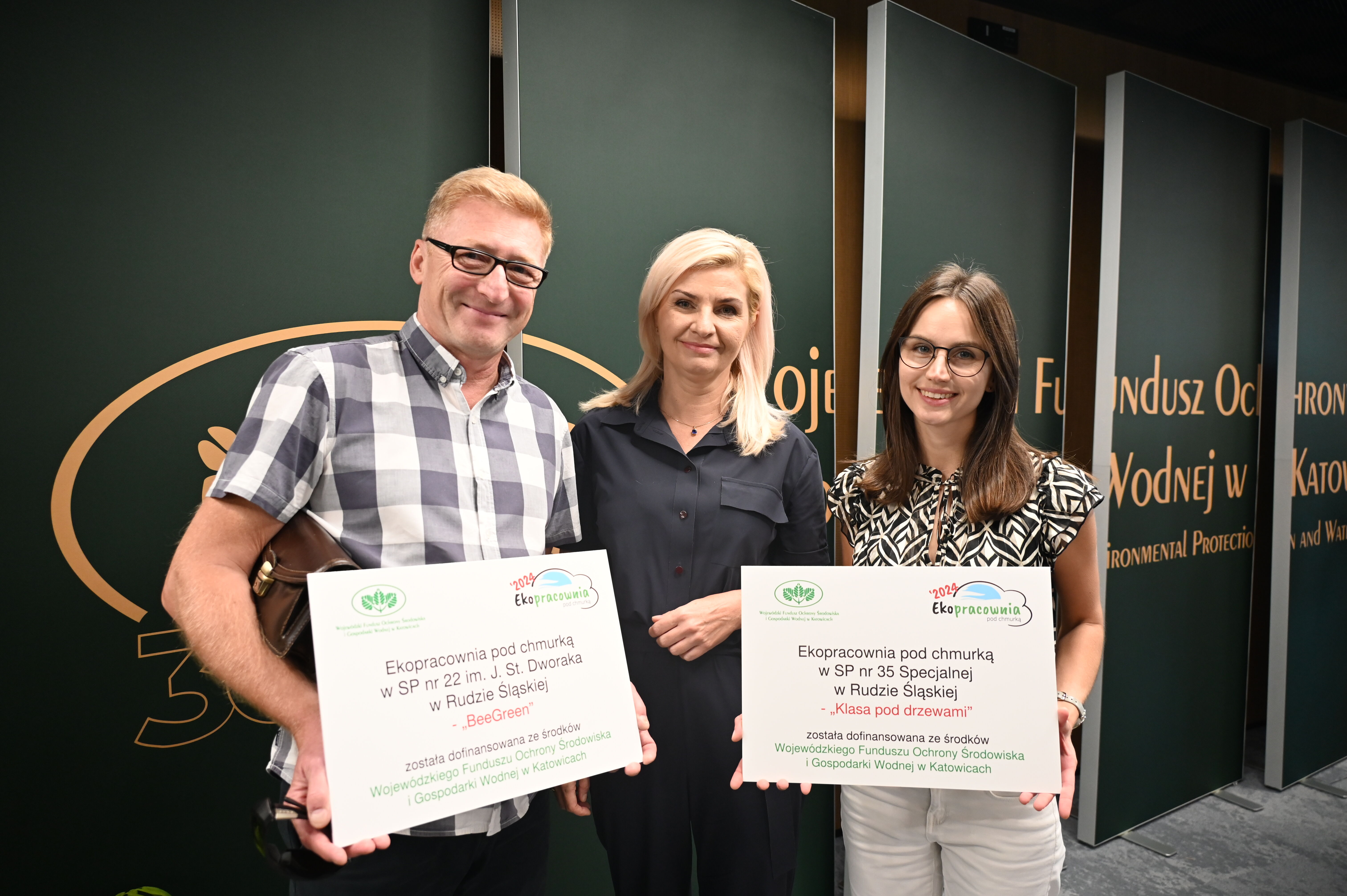 Trzy osoby - dwie kobiety i mężczyzna stoją na tle ścianki. Kobieta i mężczyzna trzymają kartonowe tabliczki.