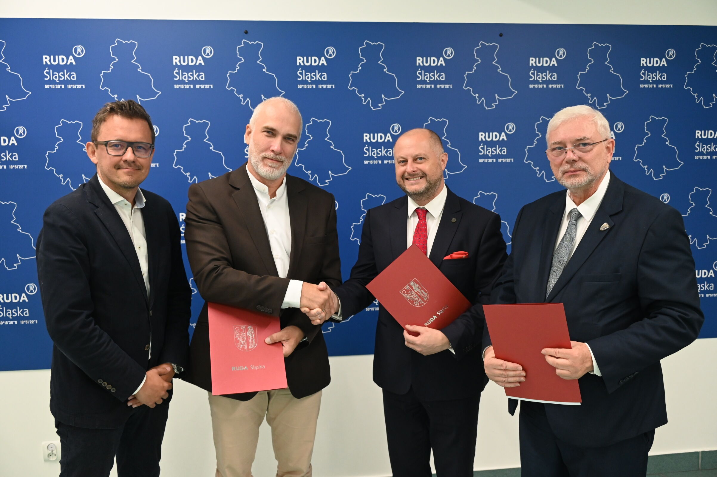Czterech mężczyzn w garniturach. Trzech trzyma teczki z herbem Rudy Śląskiej, dwóch podaje sobie ręce. W tle ścianka z herbem Rudy Śląskiej.