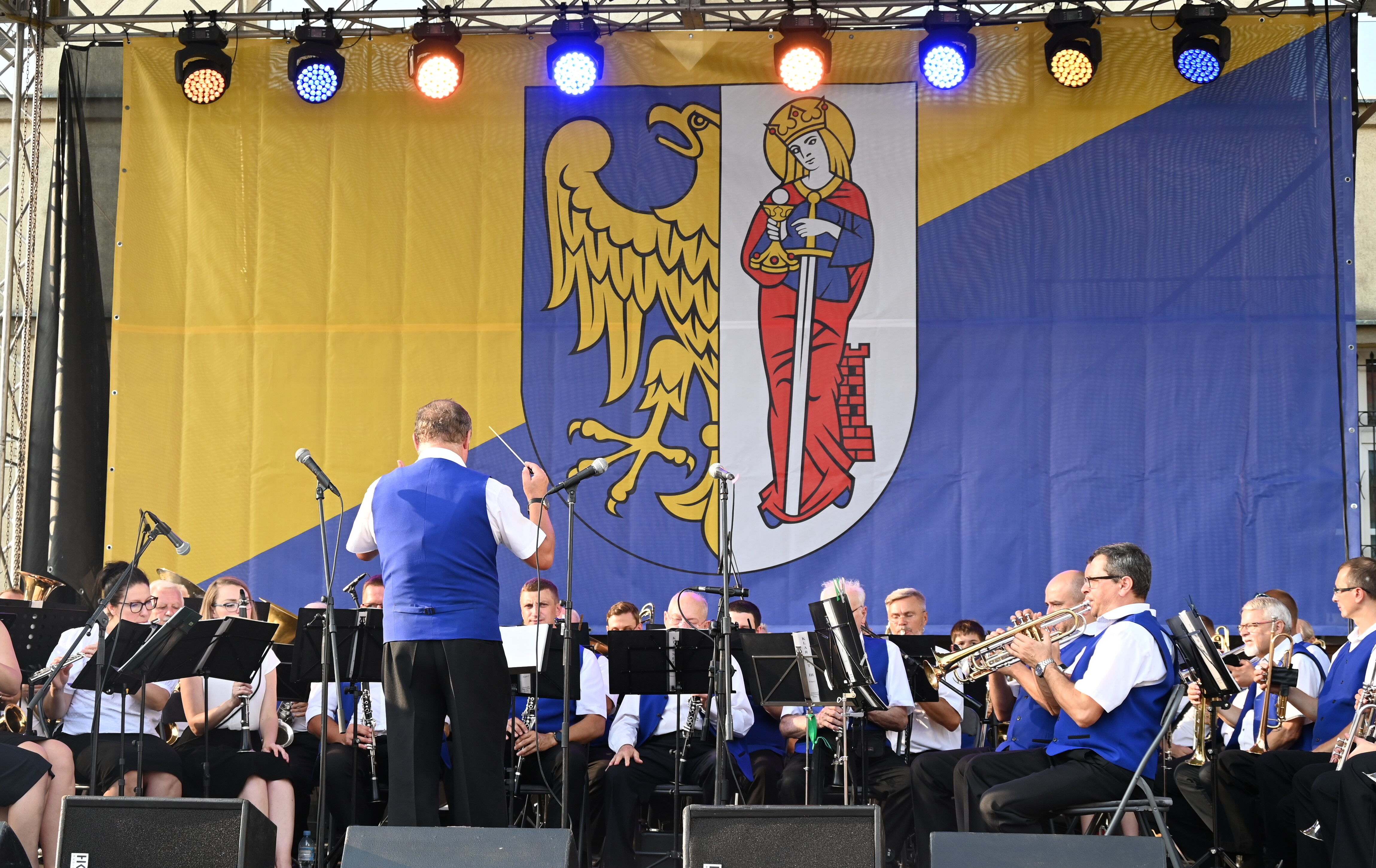 Na scenie orkiestra dęta złożona z kobiet i mężczyzn. W tle wielka flaga z herbem Rudy Śląskiej - połową orła i św. Barbarą.