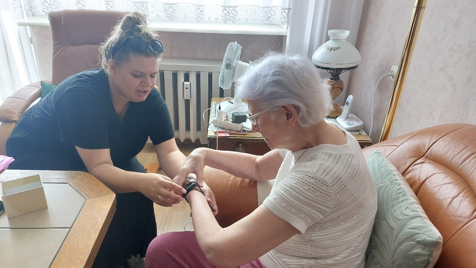 dwie kobiety siedzą przy stole, jedna pomaga drugiej założyć opaskę