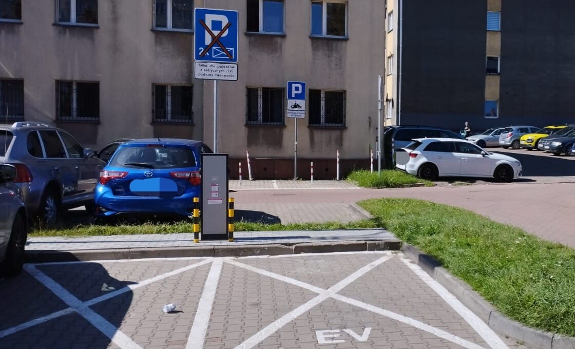 Dwa stanowiska do ładowania samochodów elektrycznych. Pionowy znak informacyjny, w tle budynek, inne zaparkowane auta.