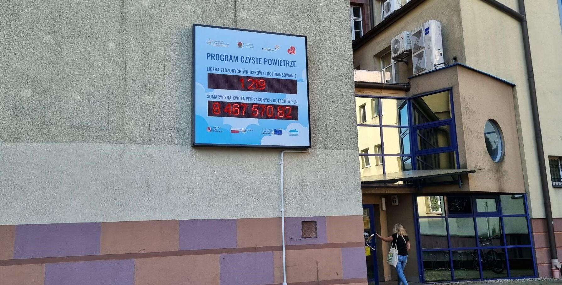 Tablica informacyjna programu "Czyste powietrze" na ścianie budynku. Na wyświetlaczu dane. Liczba złożonych wniosków: 1219. Sumaryczna kwtoa wypłaconych dotacji: 8467570,82. W tle kobieta wchodząca do budynku.